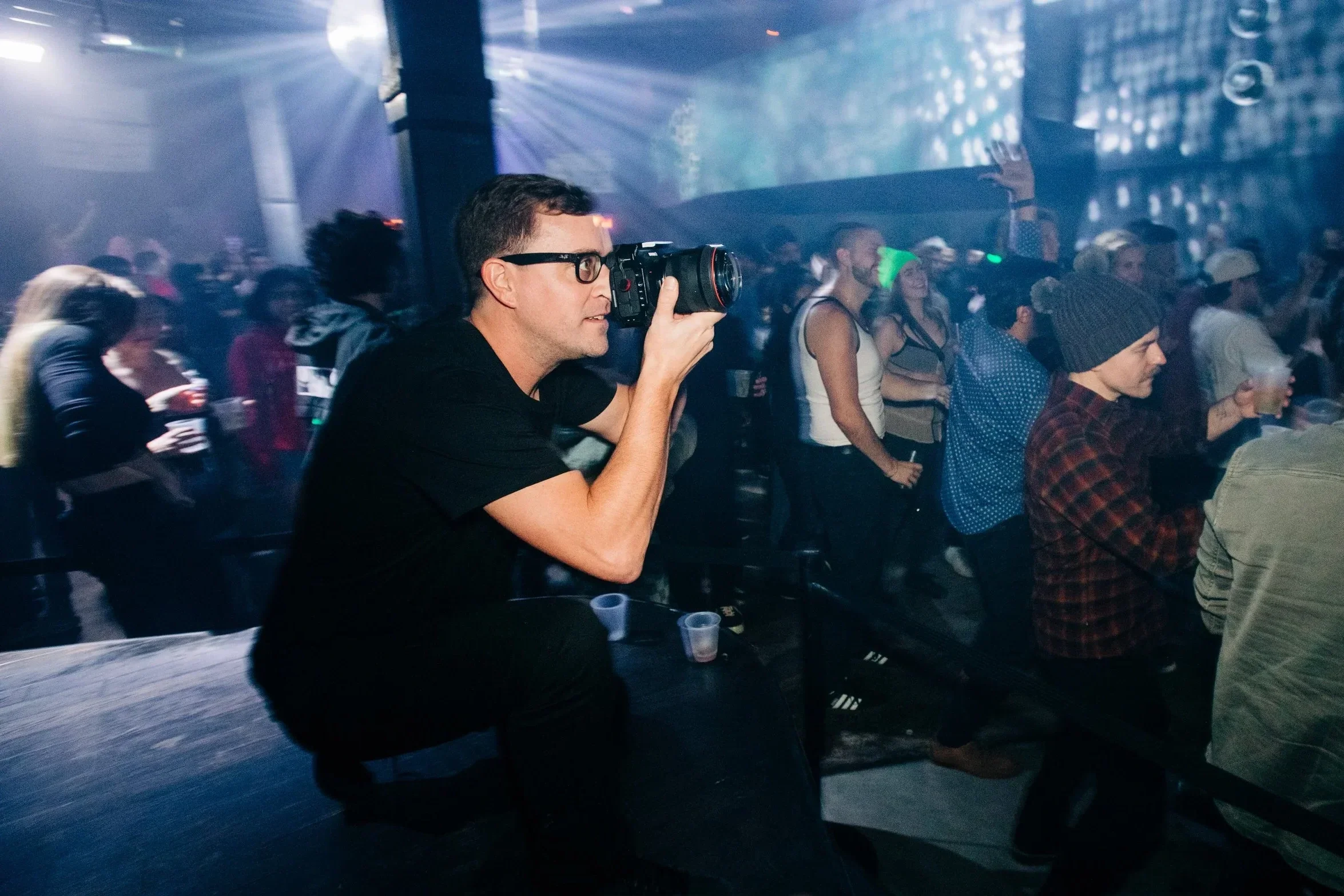A man with glasses and a black shirt taking photos with a professional camera at a crowded indoor event with colorful lights and people sitting and standing around.