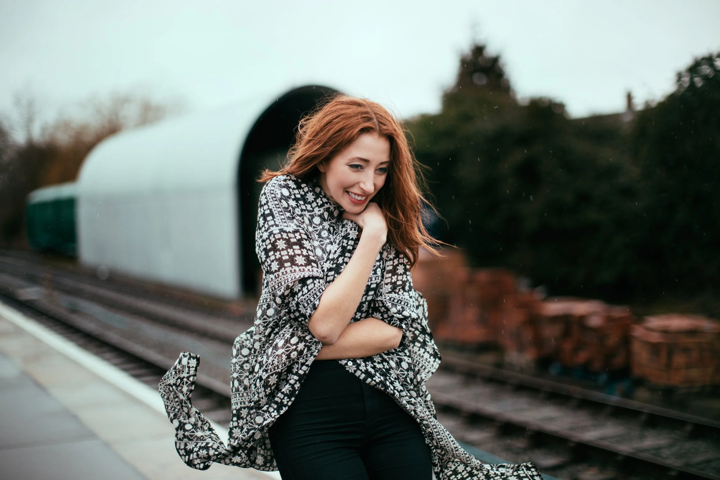 Young woman with long red hair smiling and looking down, standing outdoors near a train track, wearing a patterned jacket and black pants.