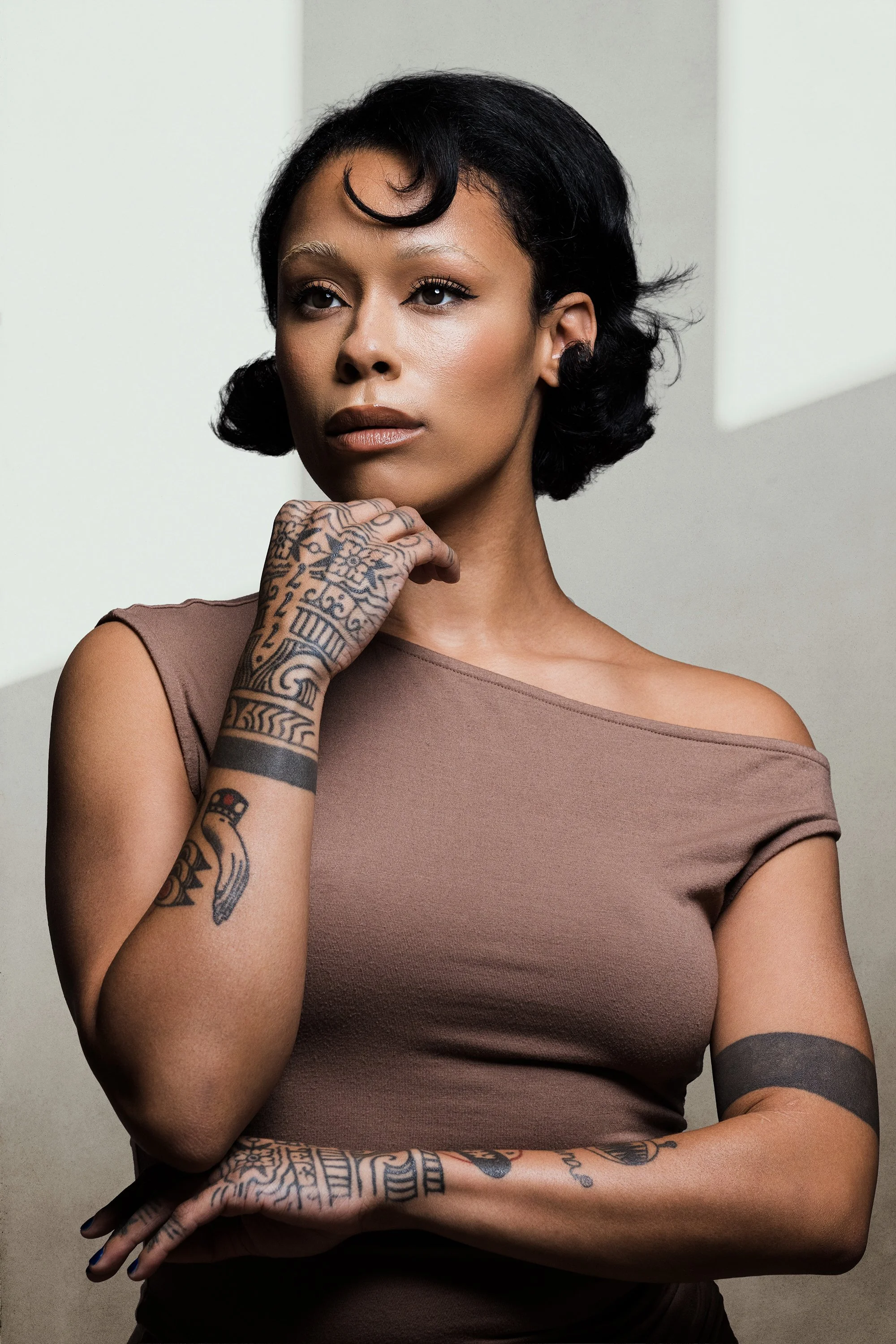 A woman with short black hair, tattoos on her right arm, and wearing a taupe-colored shirt, posing with her hand near her chin against a neutral background.