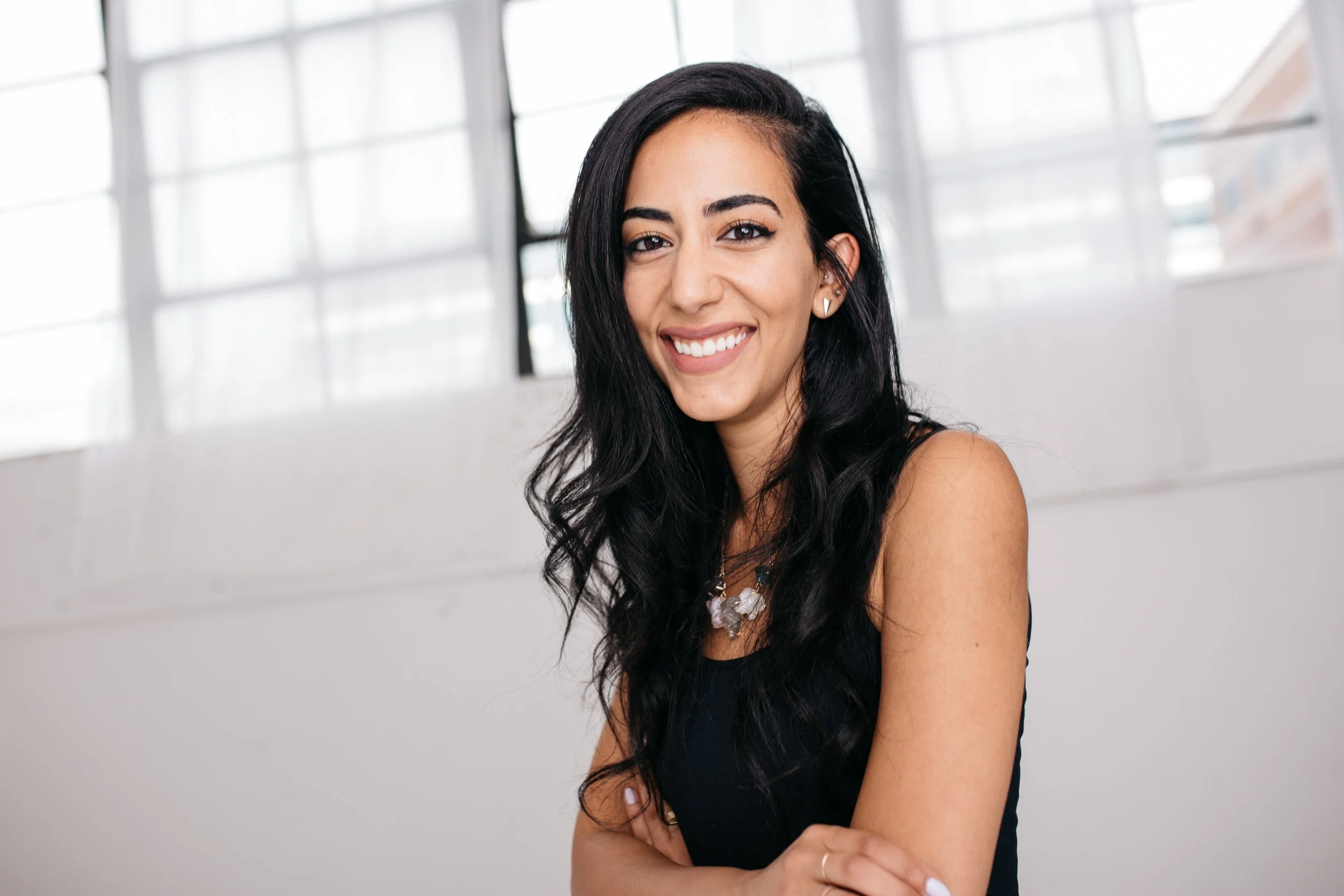 A young woman with long dark hair and a bright smile standing indoors near large windows with a white wall behind her.