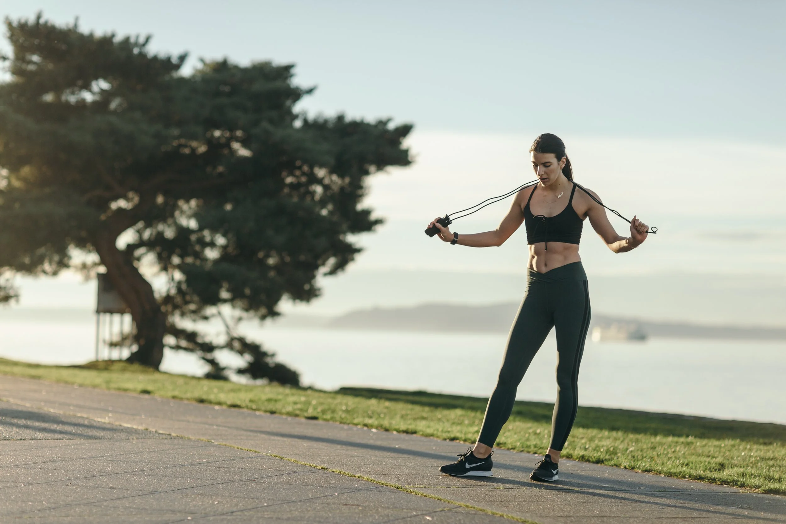 A woman outdoors, skipping rope, wearing black sports bra, black leggings, and running shoes, with trees and water in the background.