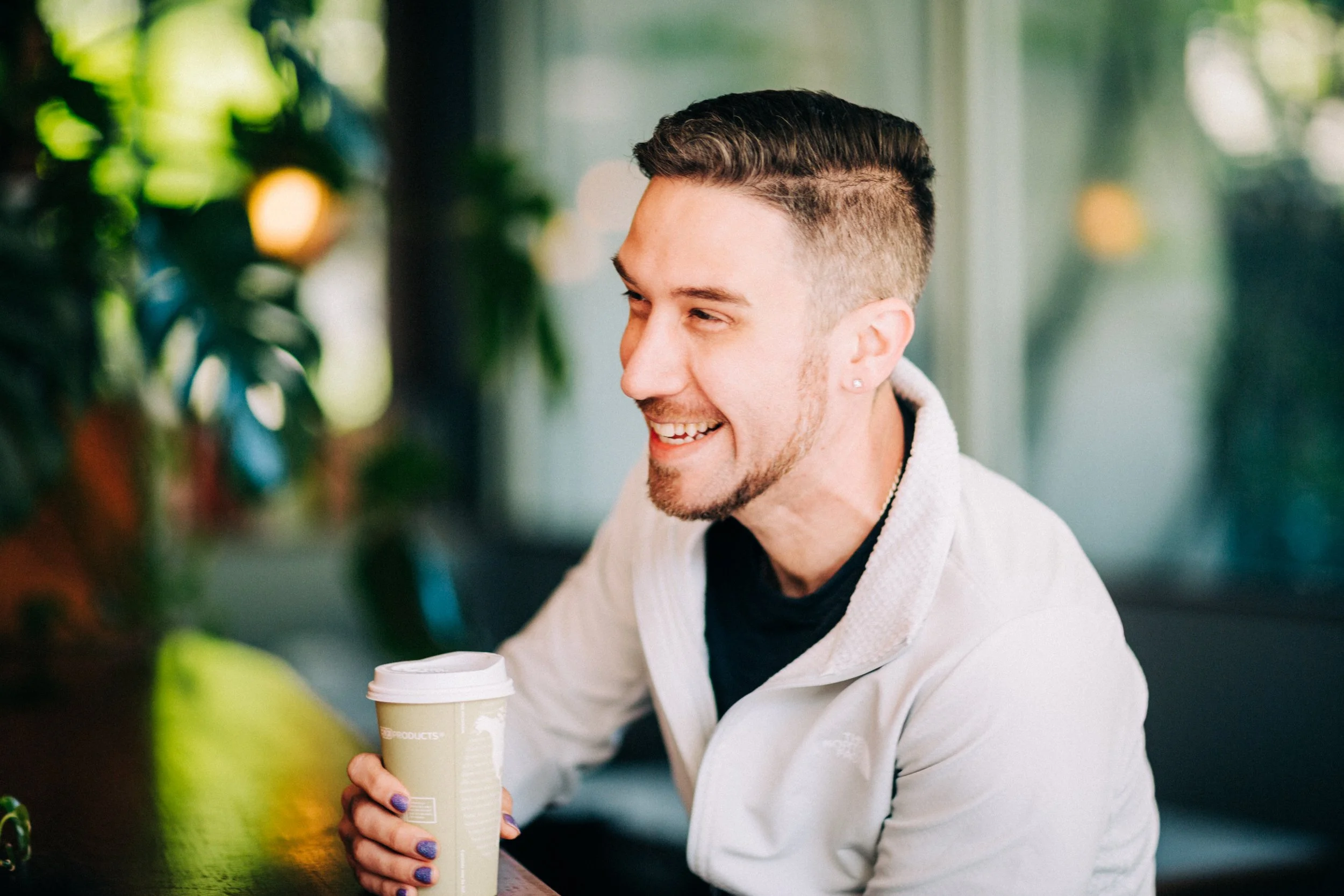 A young man with dark hair styled in undercut, smiling, holding a disposable coffee cup, inside a bright, green, plant-filled space.