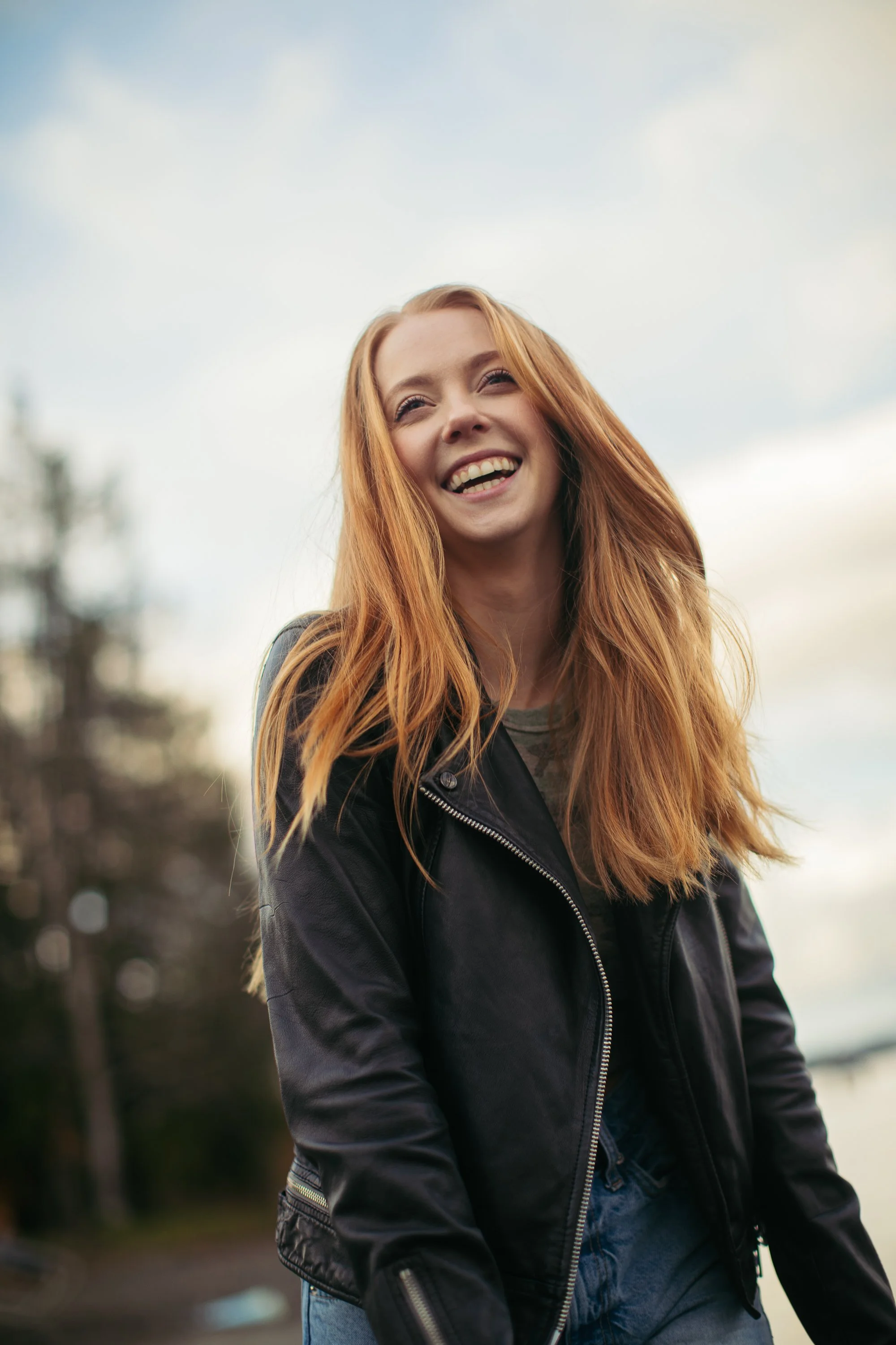 A young woman with long red hair smiling outdoors, wearing a black leather jacket and blue jeans, with a cloudy sky background.
