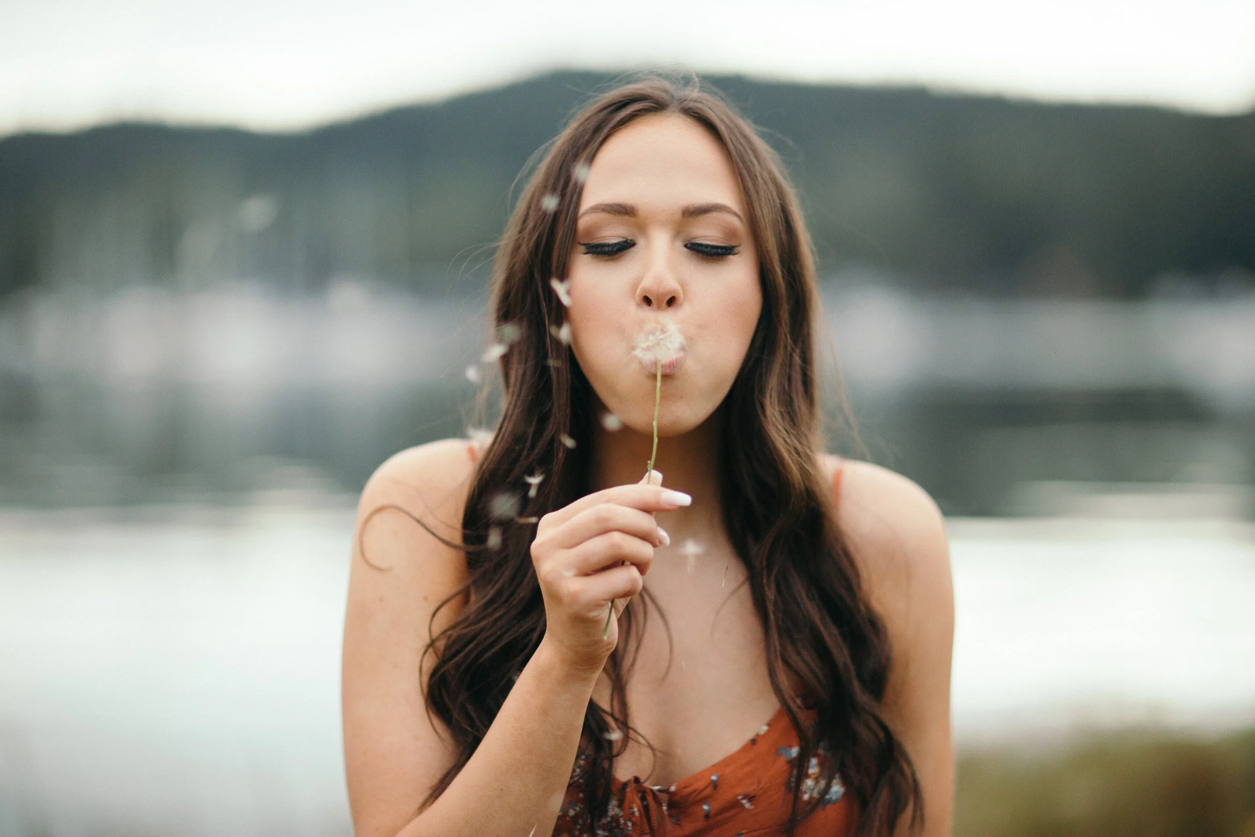 A woman with long brown hair blowing on a dandelion, with the background of a lake and mountains.