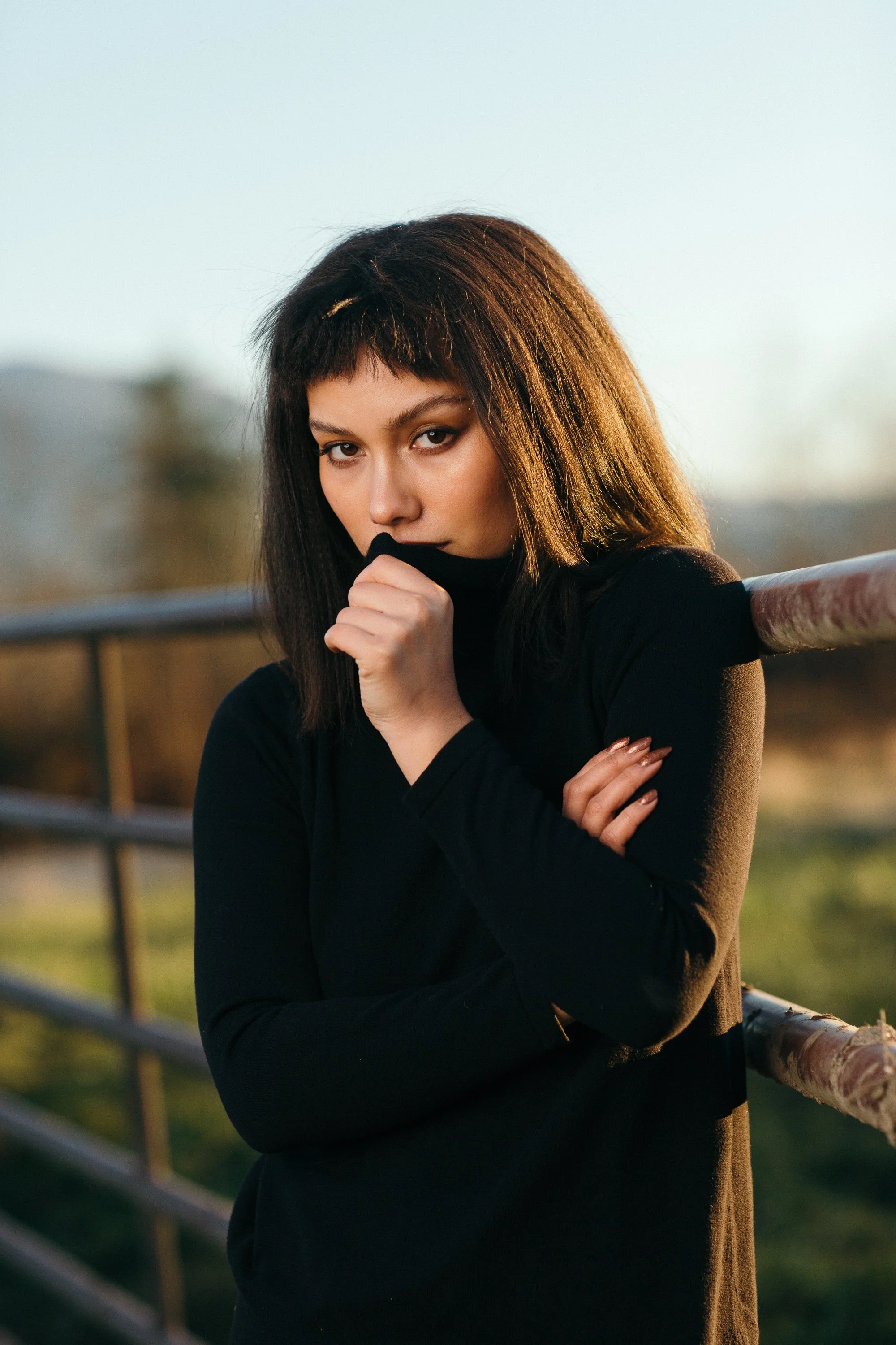 A young woman with dark brown hair and light skin, wearing a black long-sleeve shirt, is outdoors near a metal railing during sunset, with her left hand touching her face and her right arm crossed over her chest.