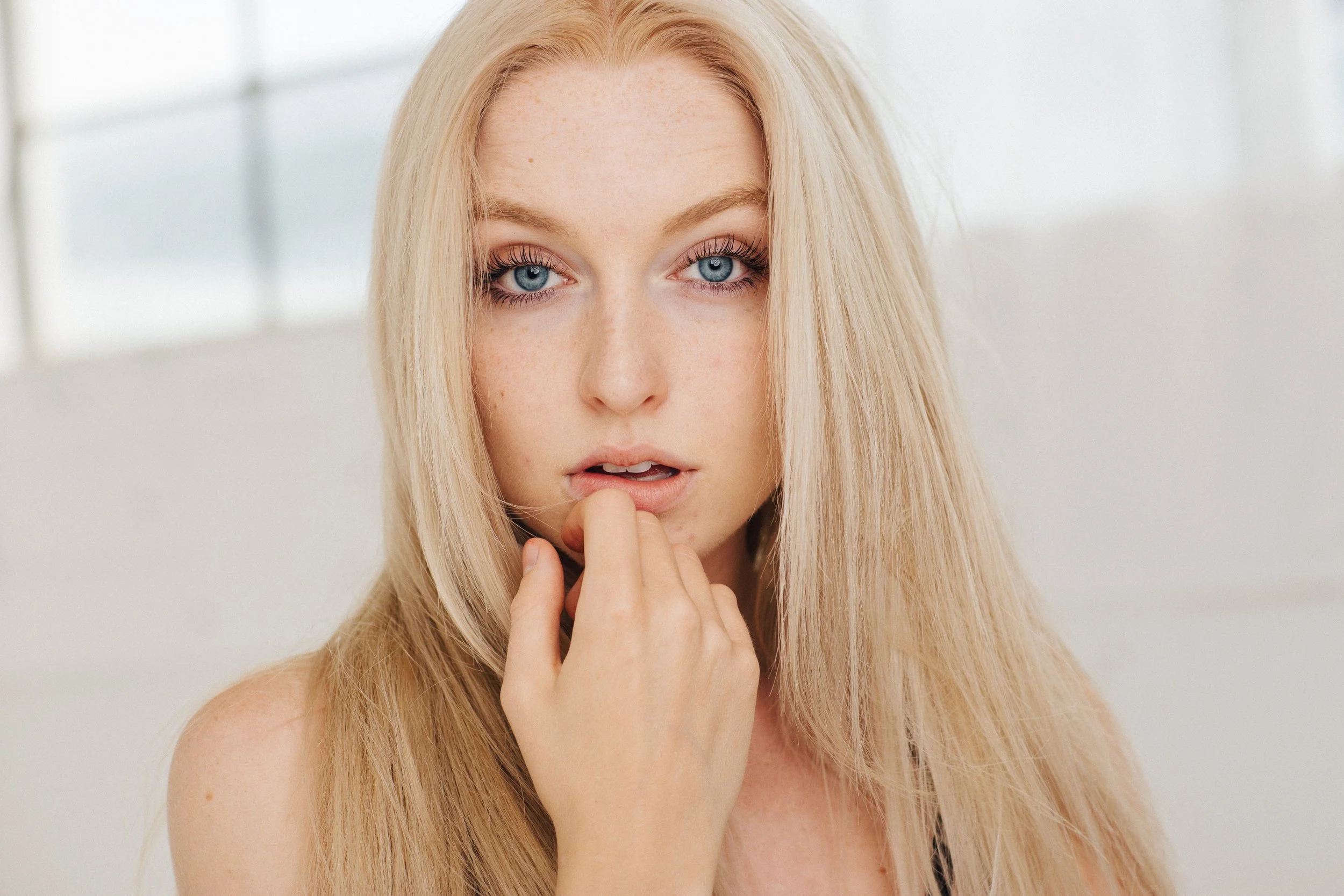 Close-up of a young woman with long blonde hair and blue eyes, touching her lips with her hand.