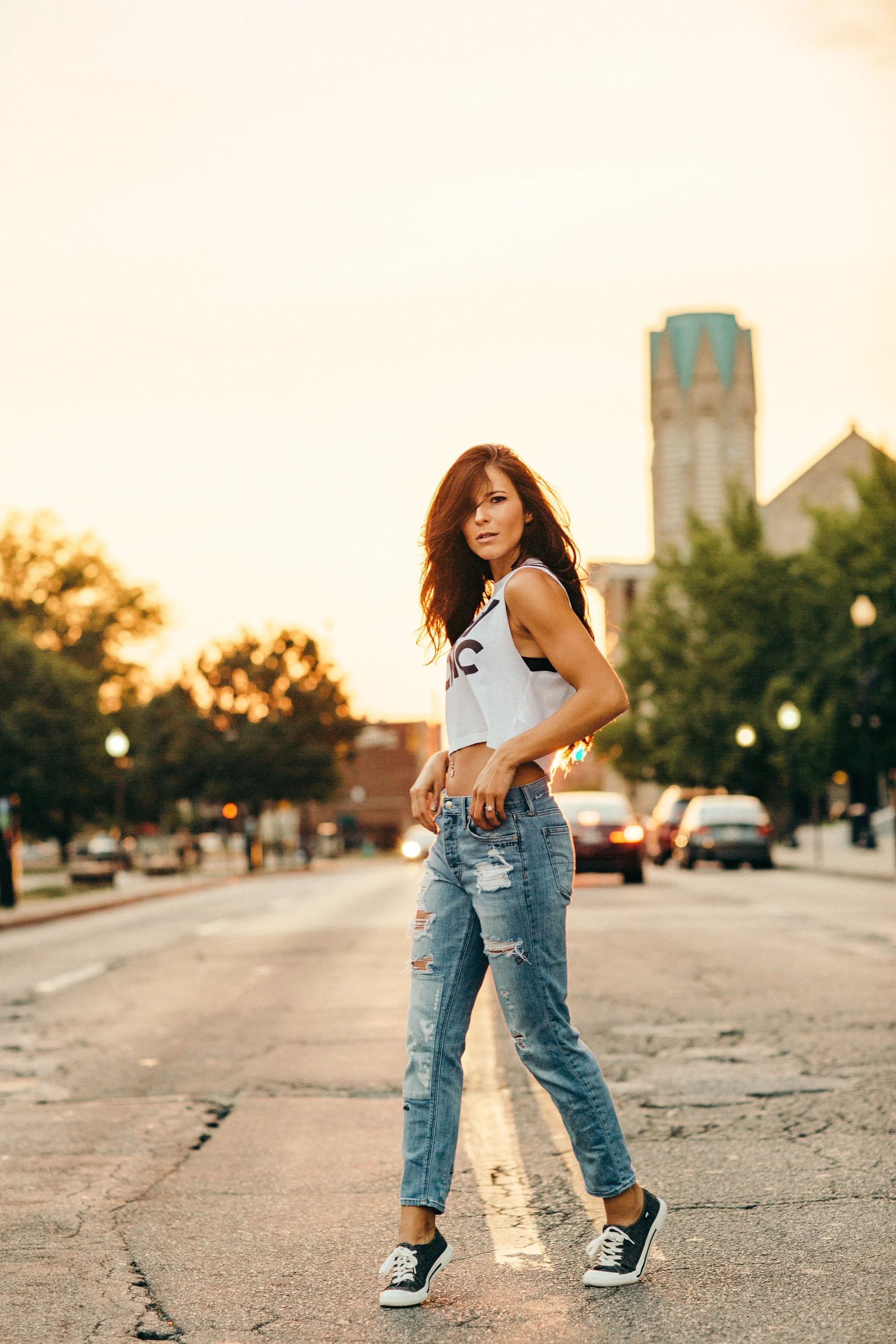 A woman with long reddish-brown hair stands in the middle of a street during sunset, wearing a white crop top with black accents and ripped blue jeans, and black and white sneakers.