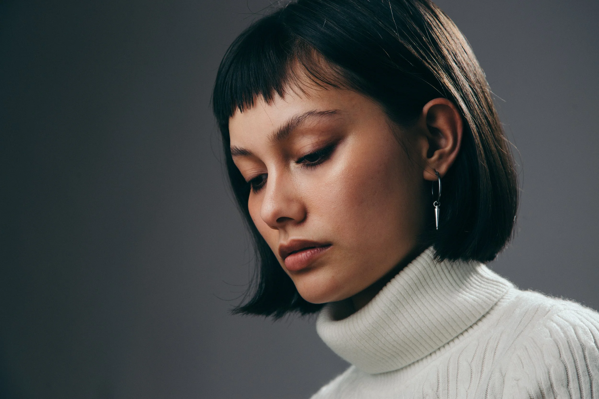 Close-up of a young woman with dark hair styled in a chin-length bob and blunt bangs, wearing a white turtleneck sweater and a dangling earring. She is looking down with a thoughtful expression, against a dark gray background.