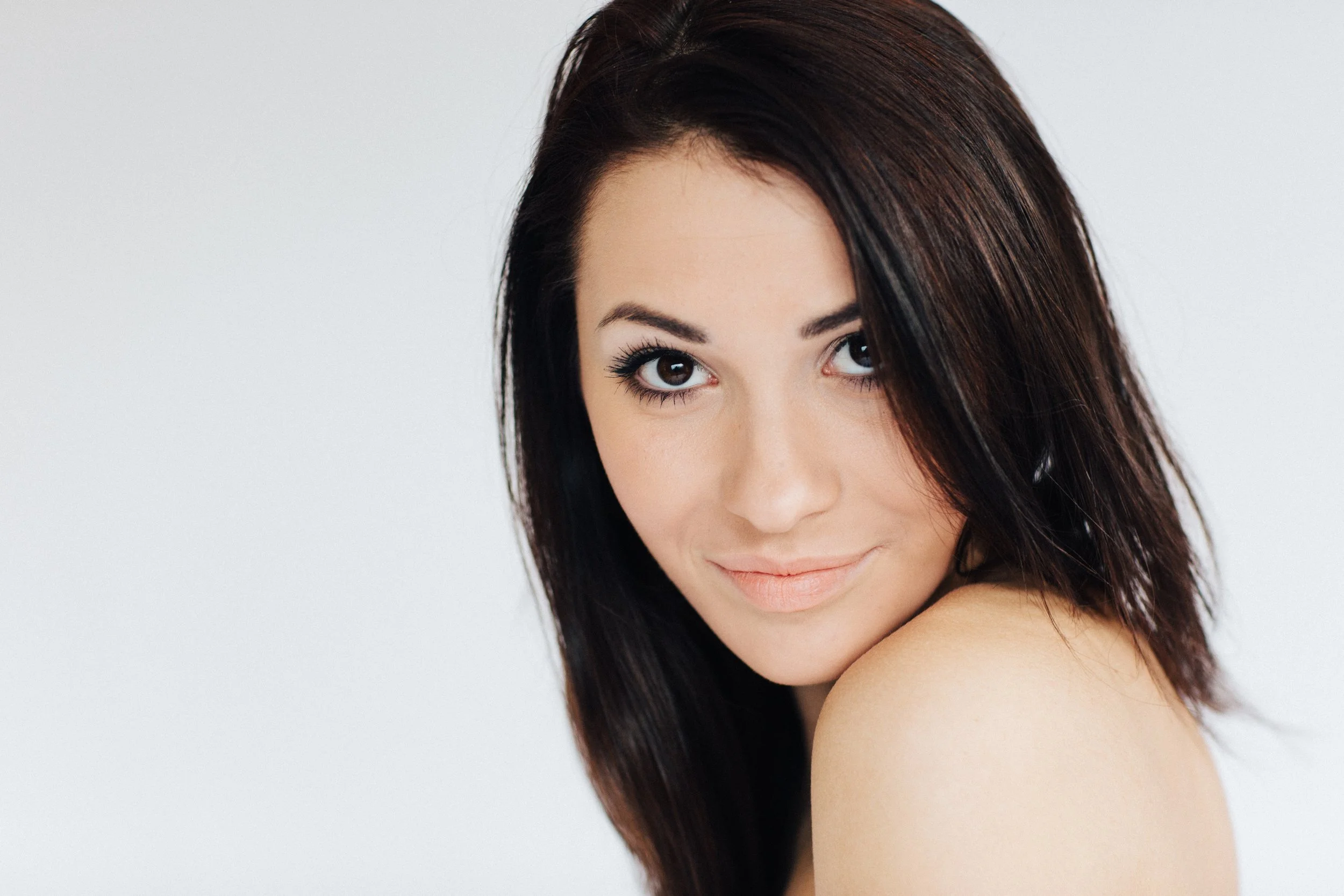 Close-up of a woman with dark brown hair, smiling softly, with a bare shoulder visible against a plain, light gray background.