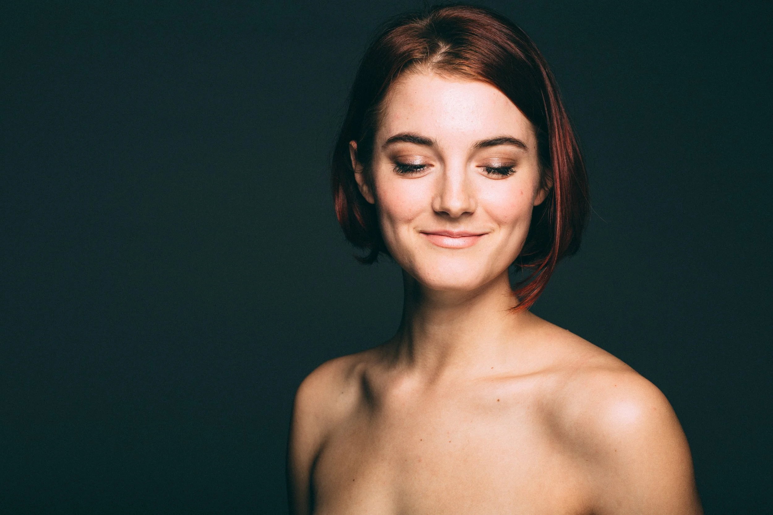 Portrait of a young woman with short brown hair, closed eyes, and a gentle smile, against a dark background.