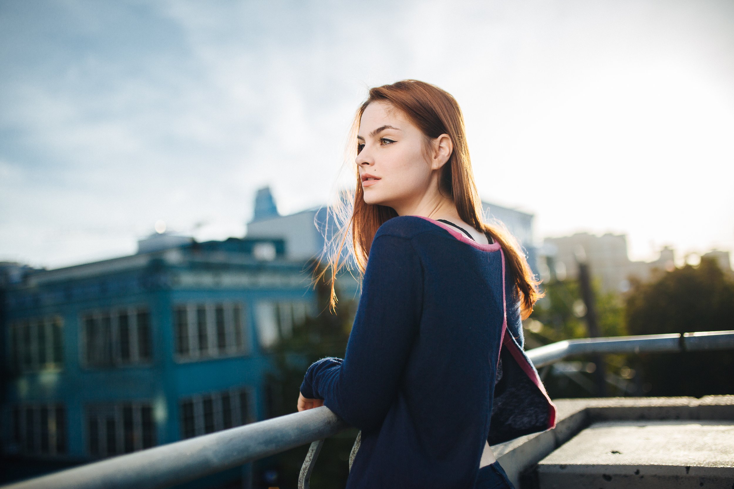 A young woman with red hair looking to the side, standing on a bridge or balcony with city buildings in the background, during late afternoon or early evening sunlight.