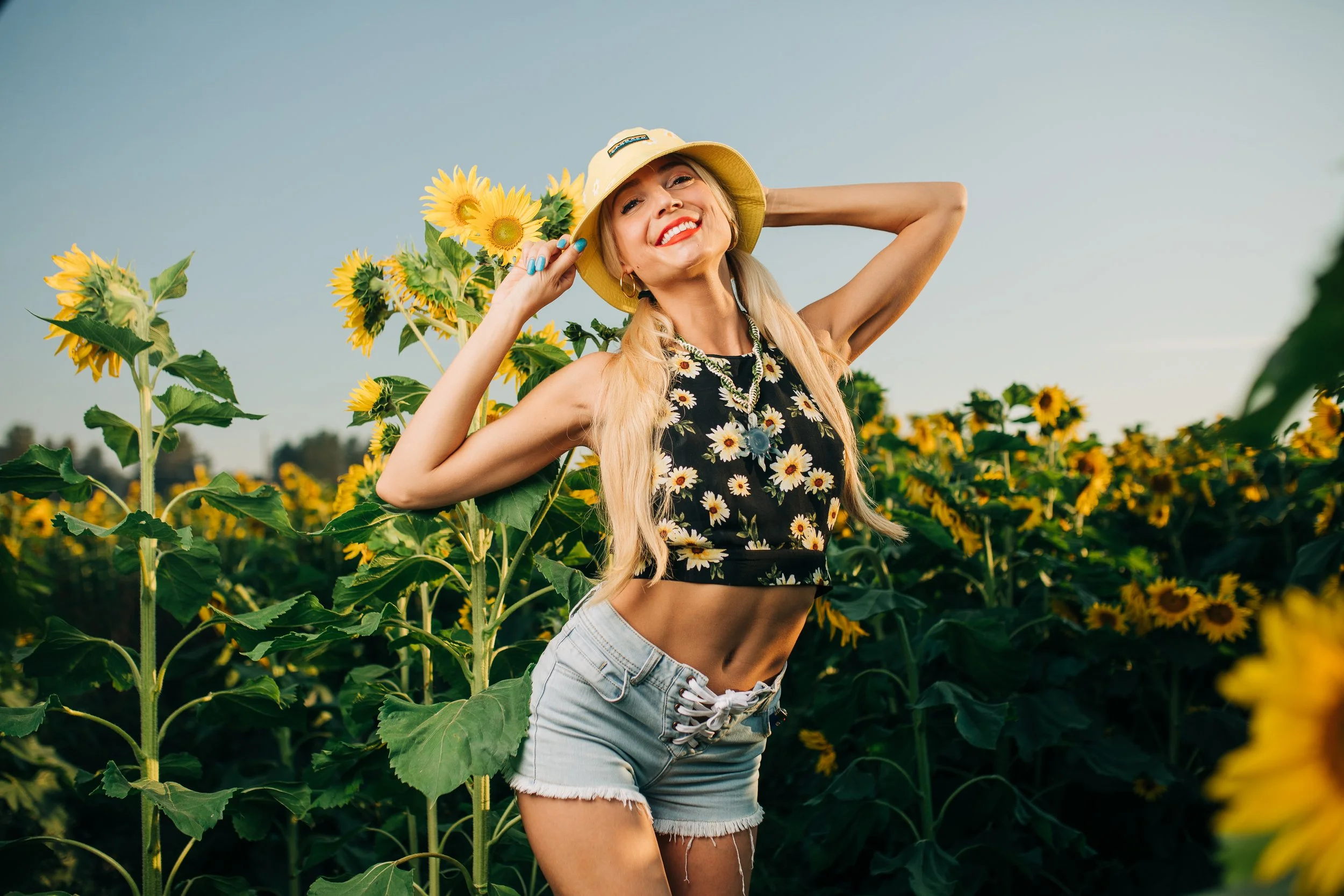 A woman with long blonde hair smiling and posing in a sunflower field, wearing a yellow hat, a black crop top with a sunflower pattern, and light denim shorts.