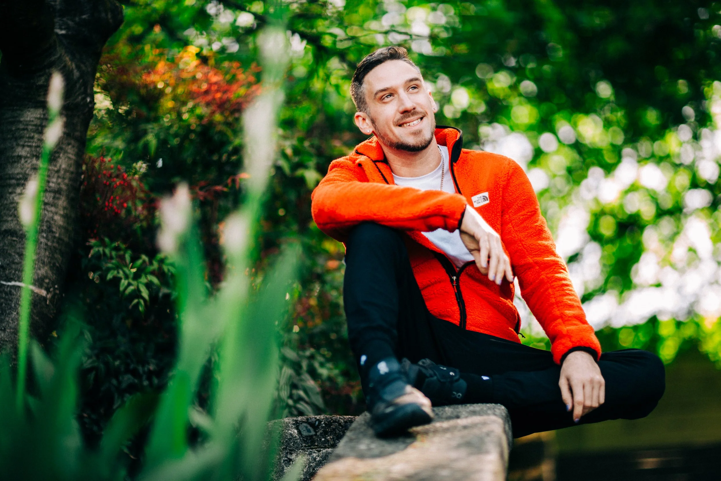 A man with short dark hair and a beard, wearing an orange jacket and black pants, sitting outdoors on a stone surface with greenery and trees in the background, smiling and looking to the side.