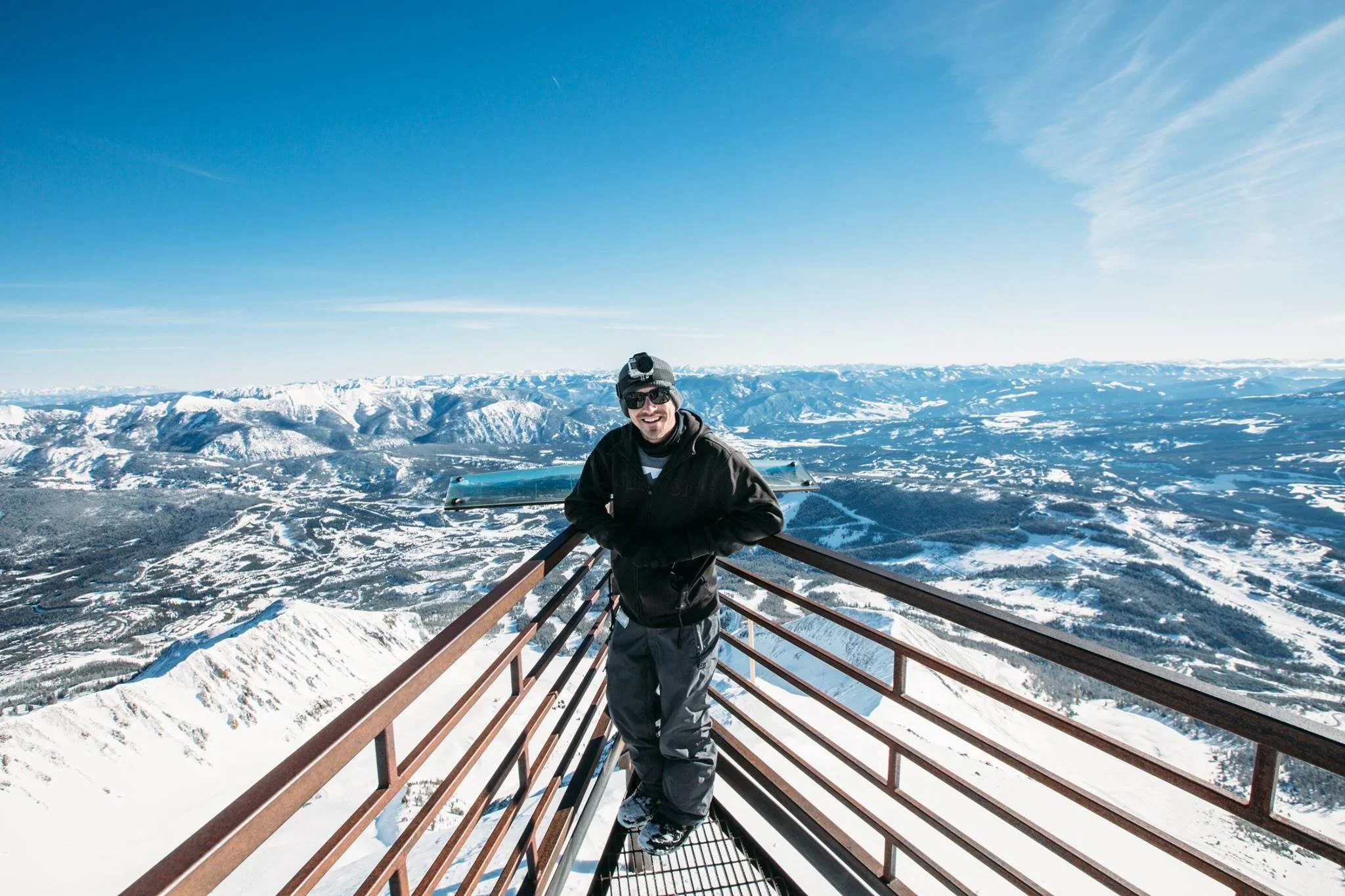Man in winter clothing with sunglasses and a headlamp, smiling, standing on a metal lookout platform overlooking snowy mountain ranges under a clear blue sky.