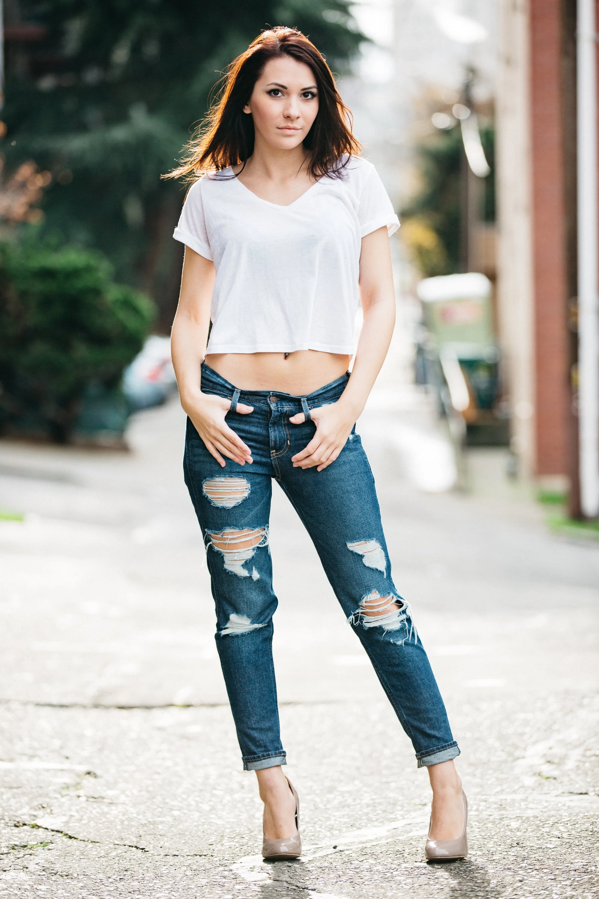 A young woman with shoulder-length dark hair standing outdoors on a street, wearing a white cropped T-shirt, distressed jeans, and beige high heels, with a confident expression.