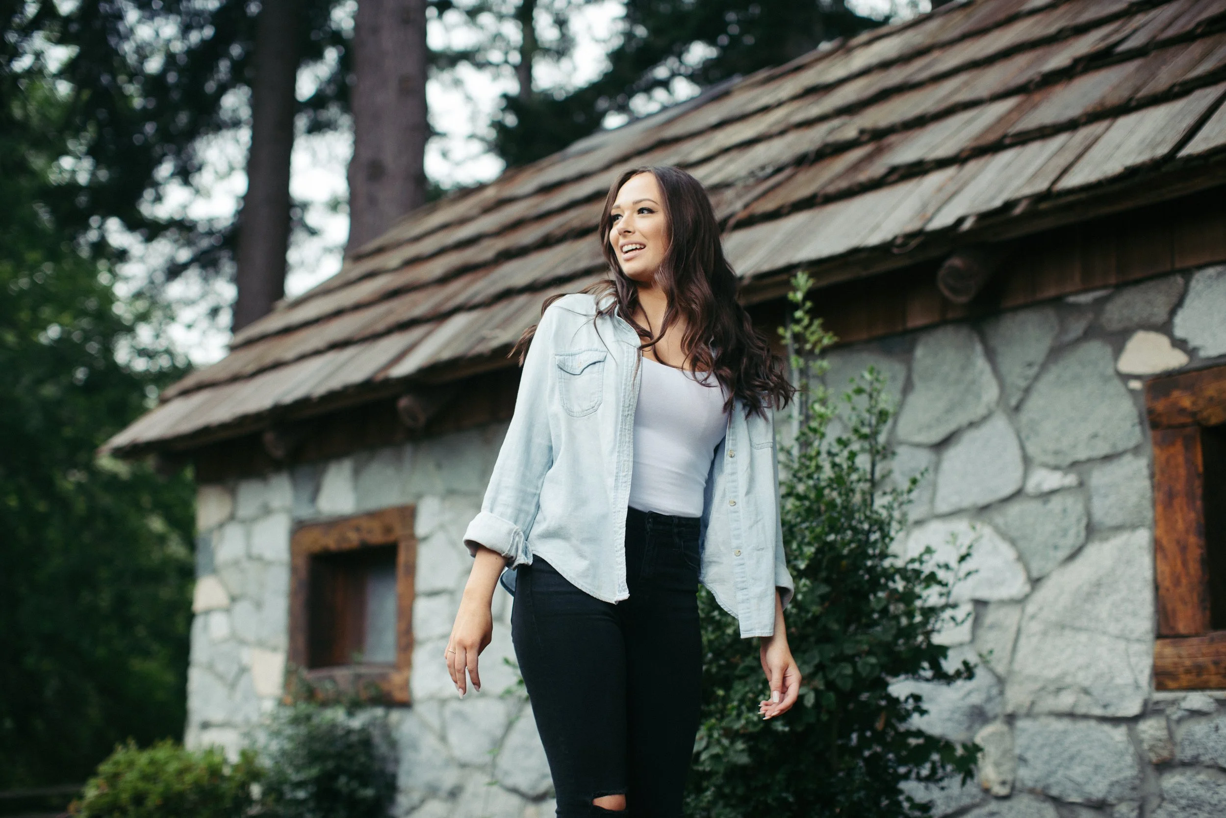 A young woman with long curly hair wearing a light denim jacket, white top, and black pants standing outdoors in front of a rustic stone house with a sloped shingled roof, surrounded by green trees and plants.