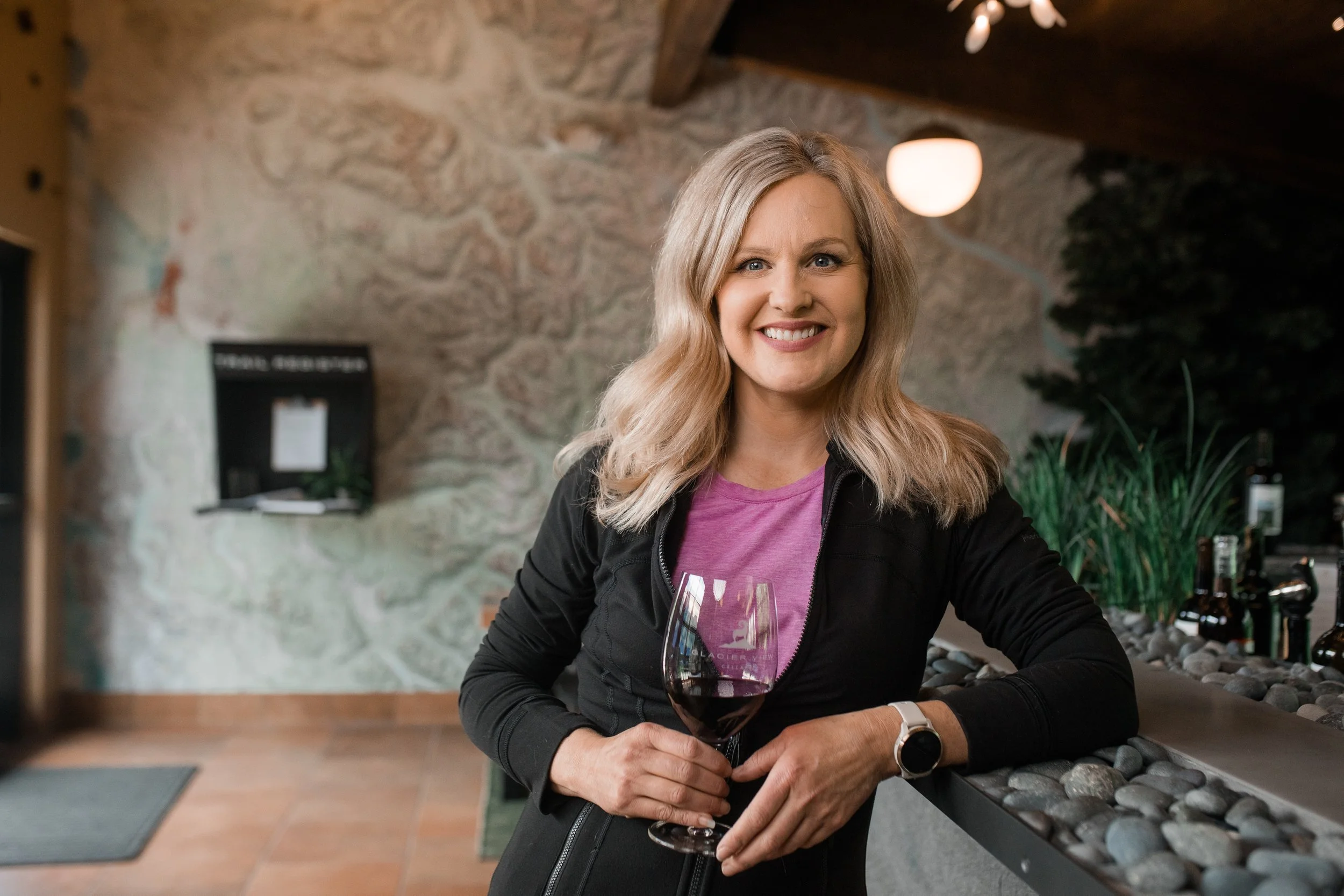 A smiling woman with blonde hair holding a glass of red wine, sitting at a bar with stone countertop. In the background, there is a stone wall, a round warm light, some potted plants, and bottles of wine.