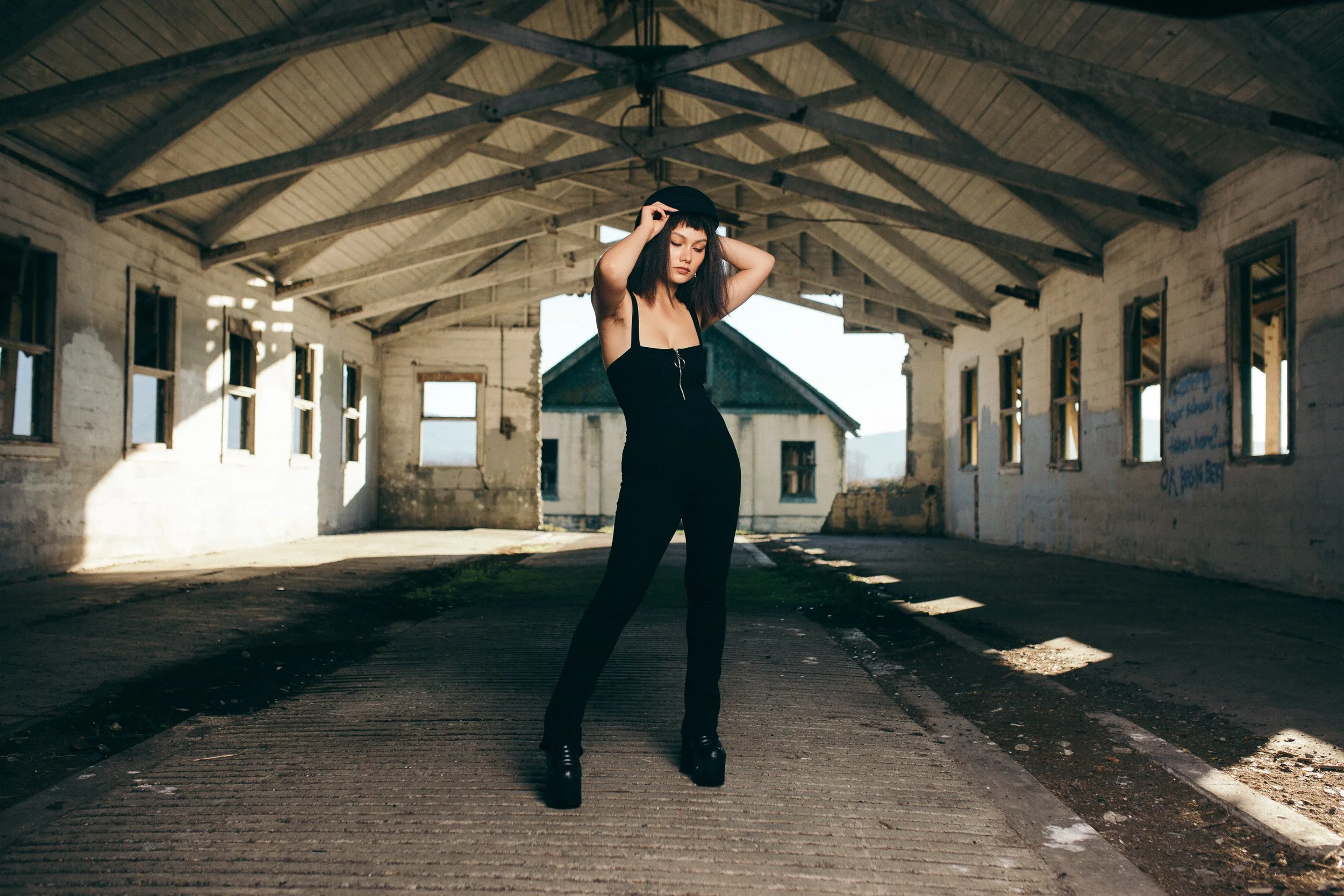 A woman in a black dress and platform boots standing under an old, abandoned building with a large, open interior and broken windows, with her eyes closed and one hand touching her head.