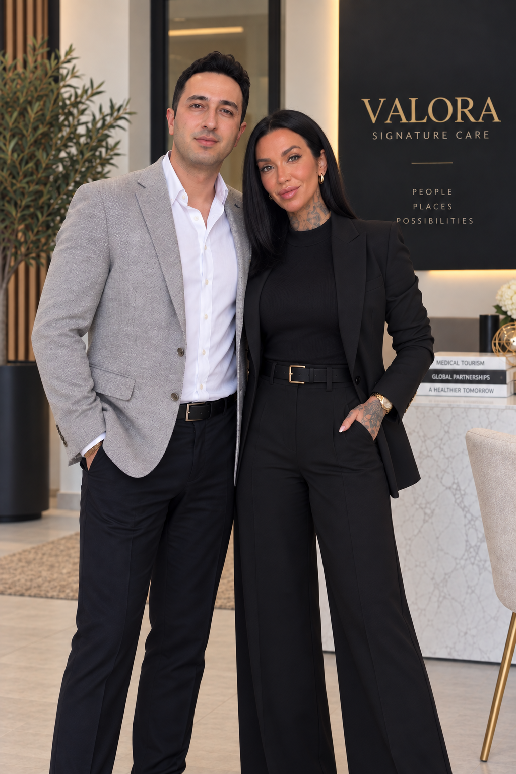 A man and a woman posing together indoors, dressed in business attire, standing in front of a sign that reads 'VALORA SIGNATURE CARE' and other promotional text.
