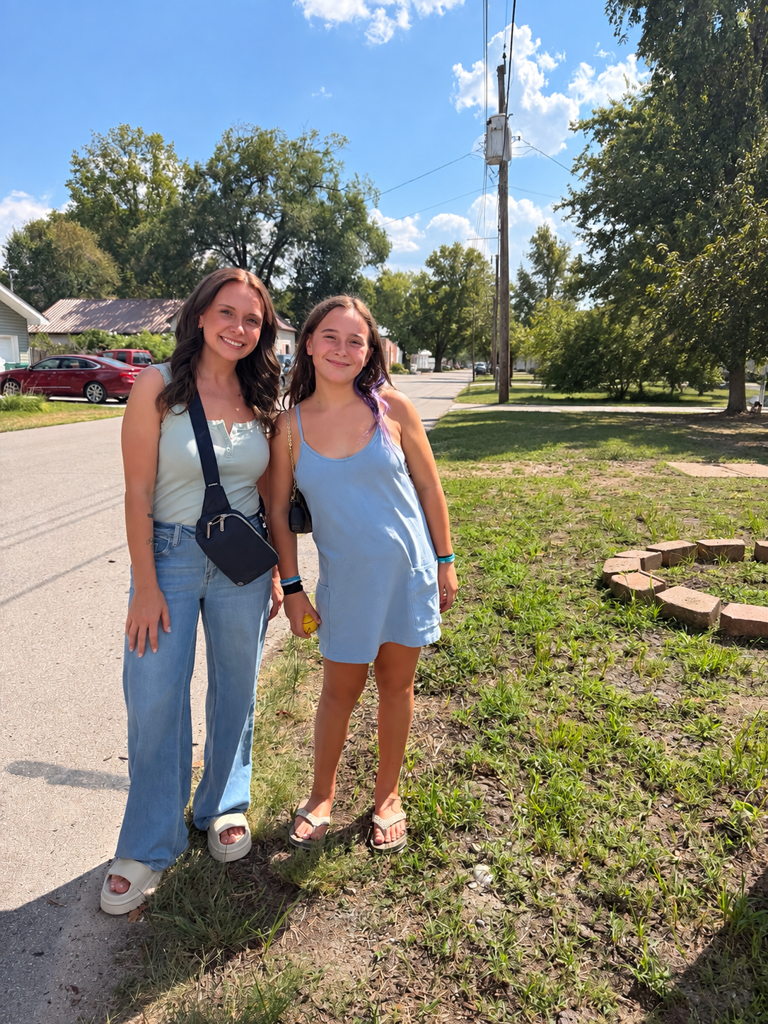 Two young women standing outdoors on a sunny day, smiling, with one wearing a light blue dress and the other wearing jeans and a sleeveless top, near a grassy area and trees.