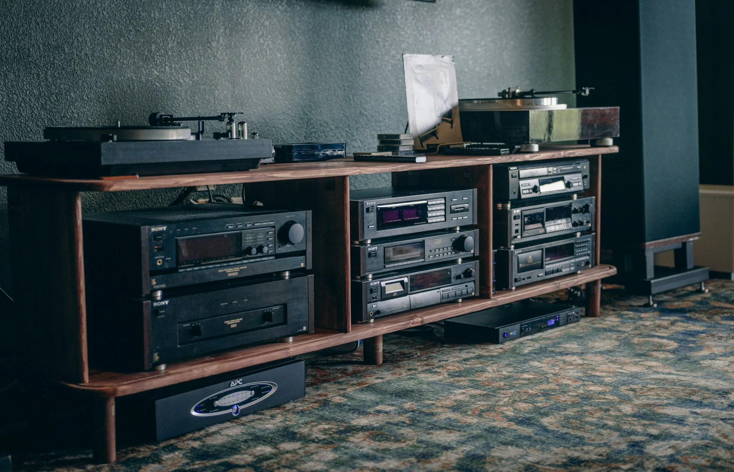 A wooden media console holding two turntables, multiple stereo receivers, cassette decks, and other audio equipment, with a dark textured wall behind and a patterned area rug on the floor.