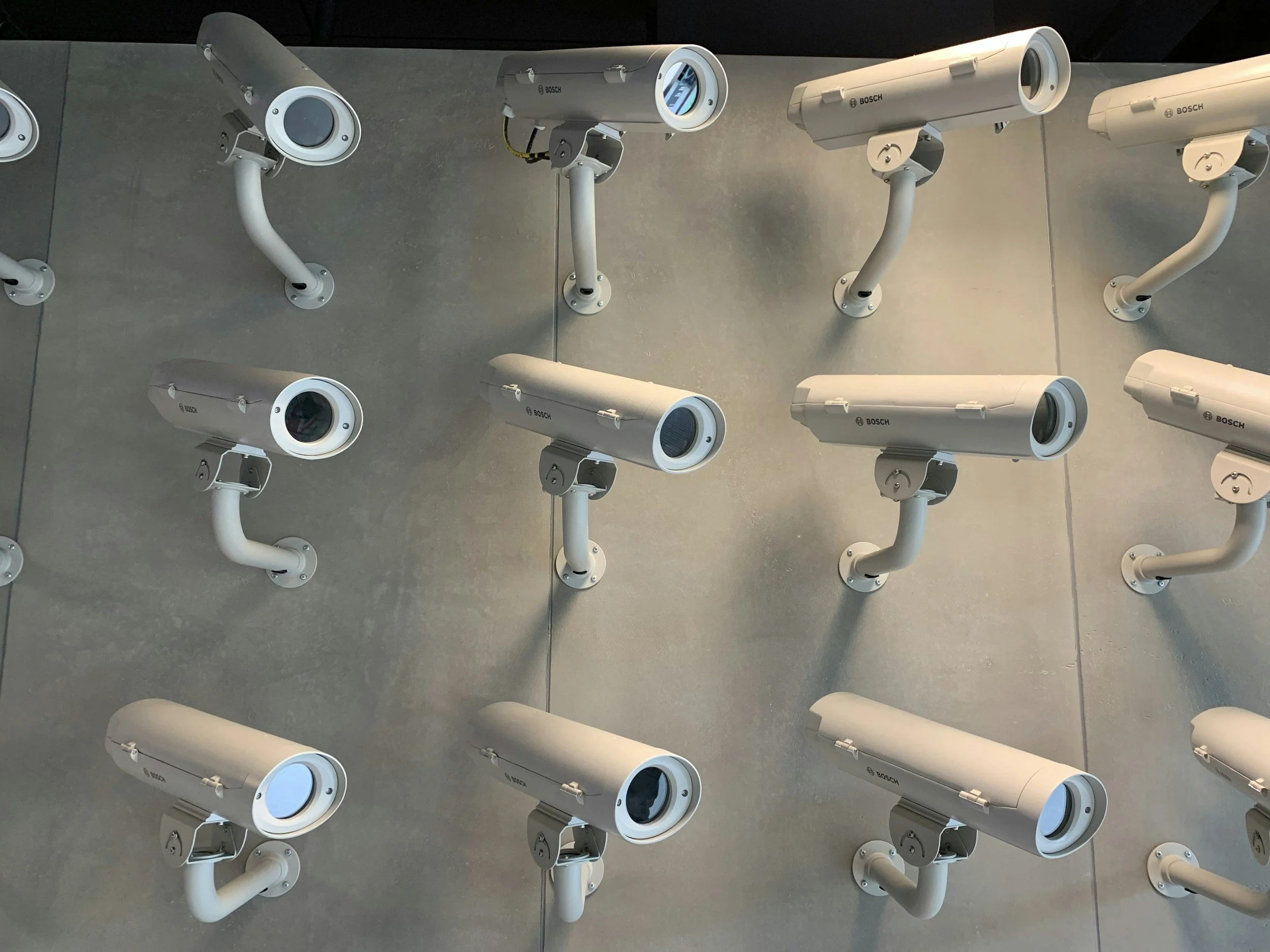 Multiple white security cameras mounted on a gray wall, with some cameras facing downward and others facing upward.