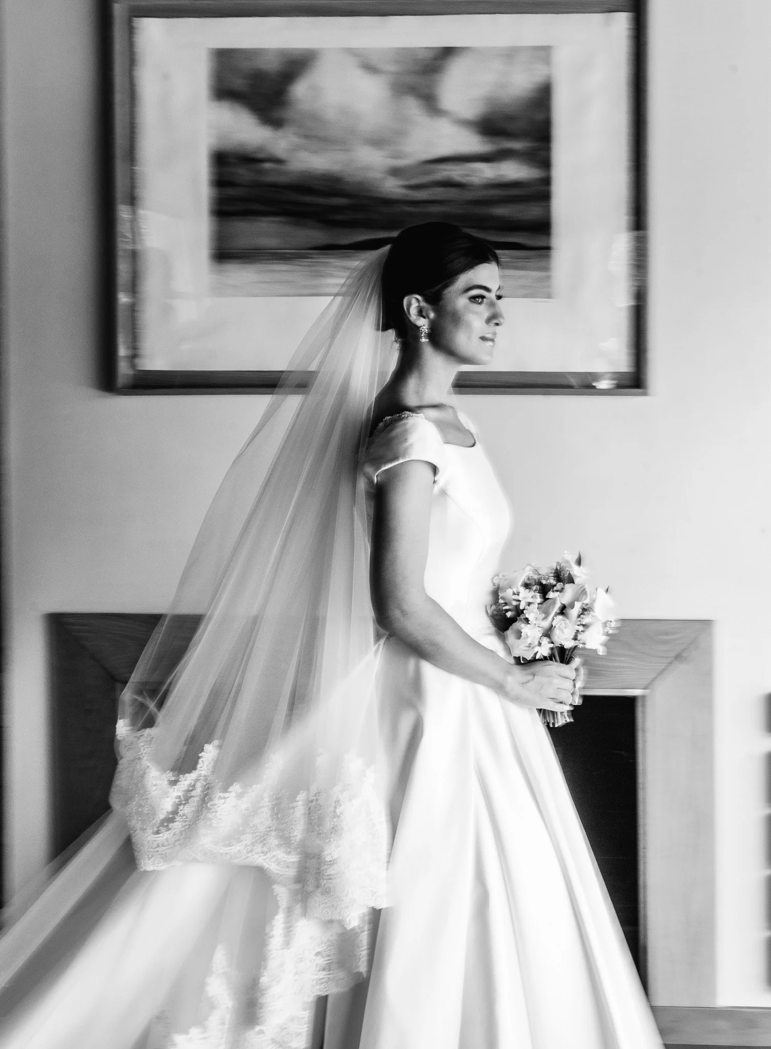 Une mariée en robe blanche tenant un bouquet de fleurs, regardant vers la droite, avec un voile, dans une pièce avec un tableau accroché au mur.