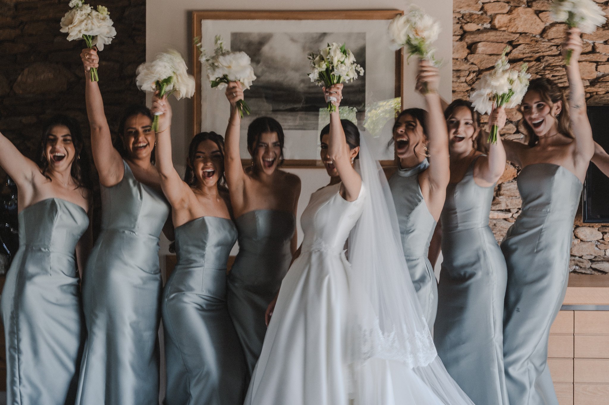 Un groupe de femmes habillées en robes de mariée, souriantes, levant leurs bouquets de fleurs blanches dans une pièce avec un mur en pierre.