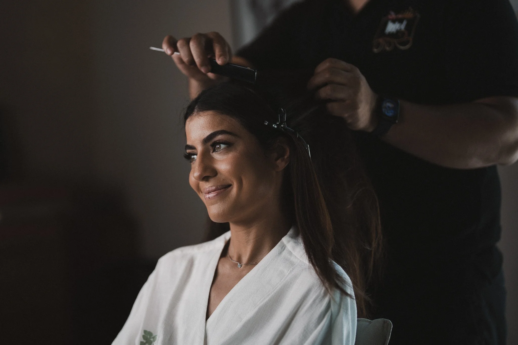 Une femme souriante est en cours de coiffure, une personne manipule ses cheveux avec des outils de coiffure dans une ambiance sombre.