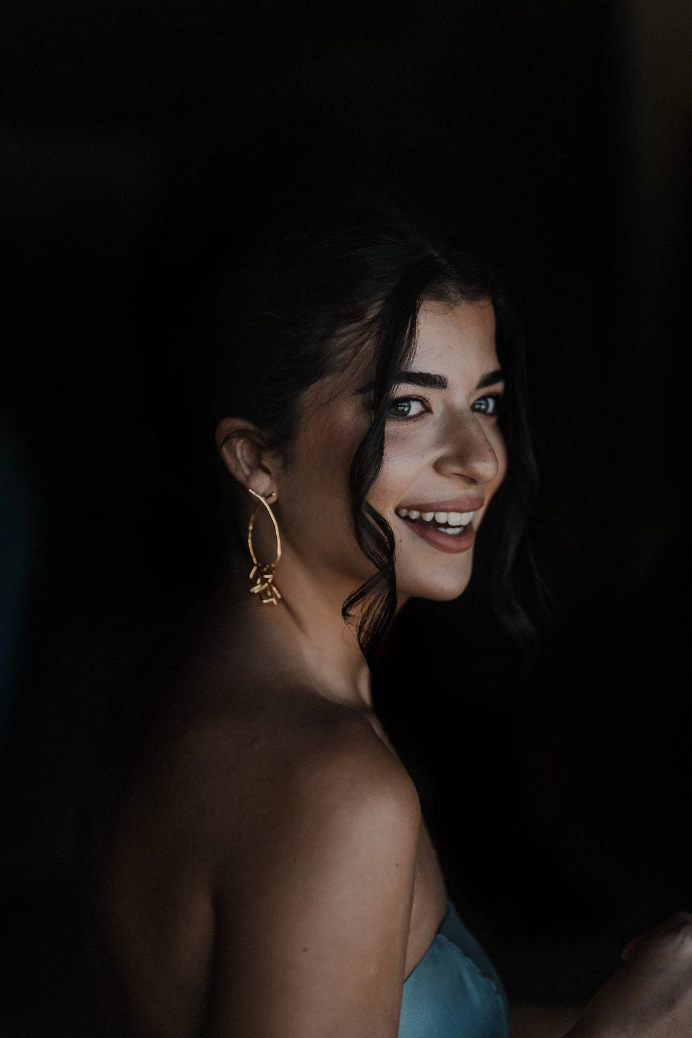 Une femme souriante avec des cheveux noirs, portant de grandes boucles d'oreilles dorées, dans un éclairage faible, portant un haut bleu.