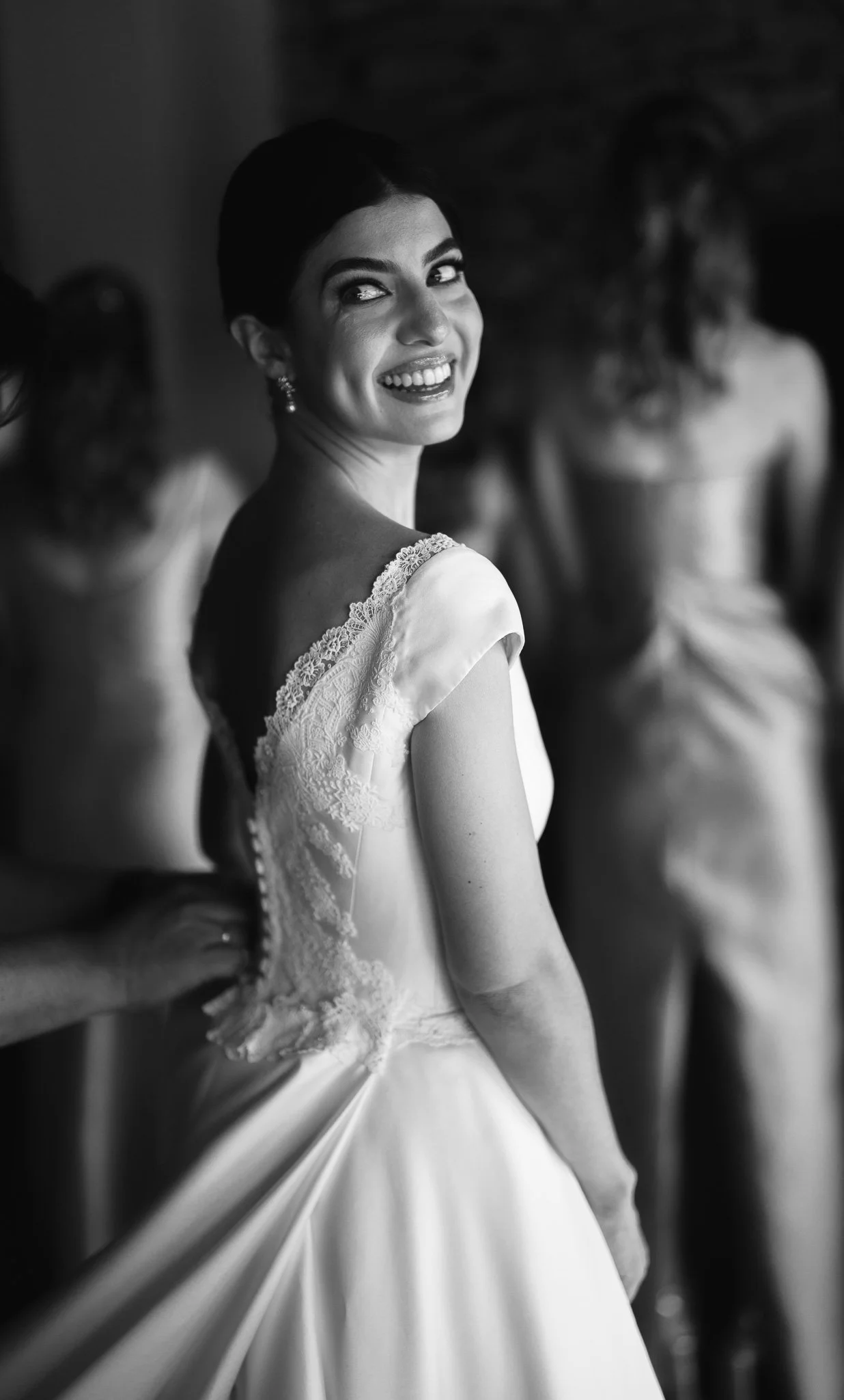 Une femme souriante en robe de mariée, regardant par-dessus son épaule, avec d'autres femmes en robes de mariée floues en arrière-plan.