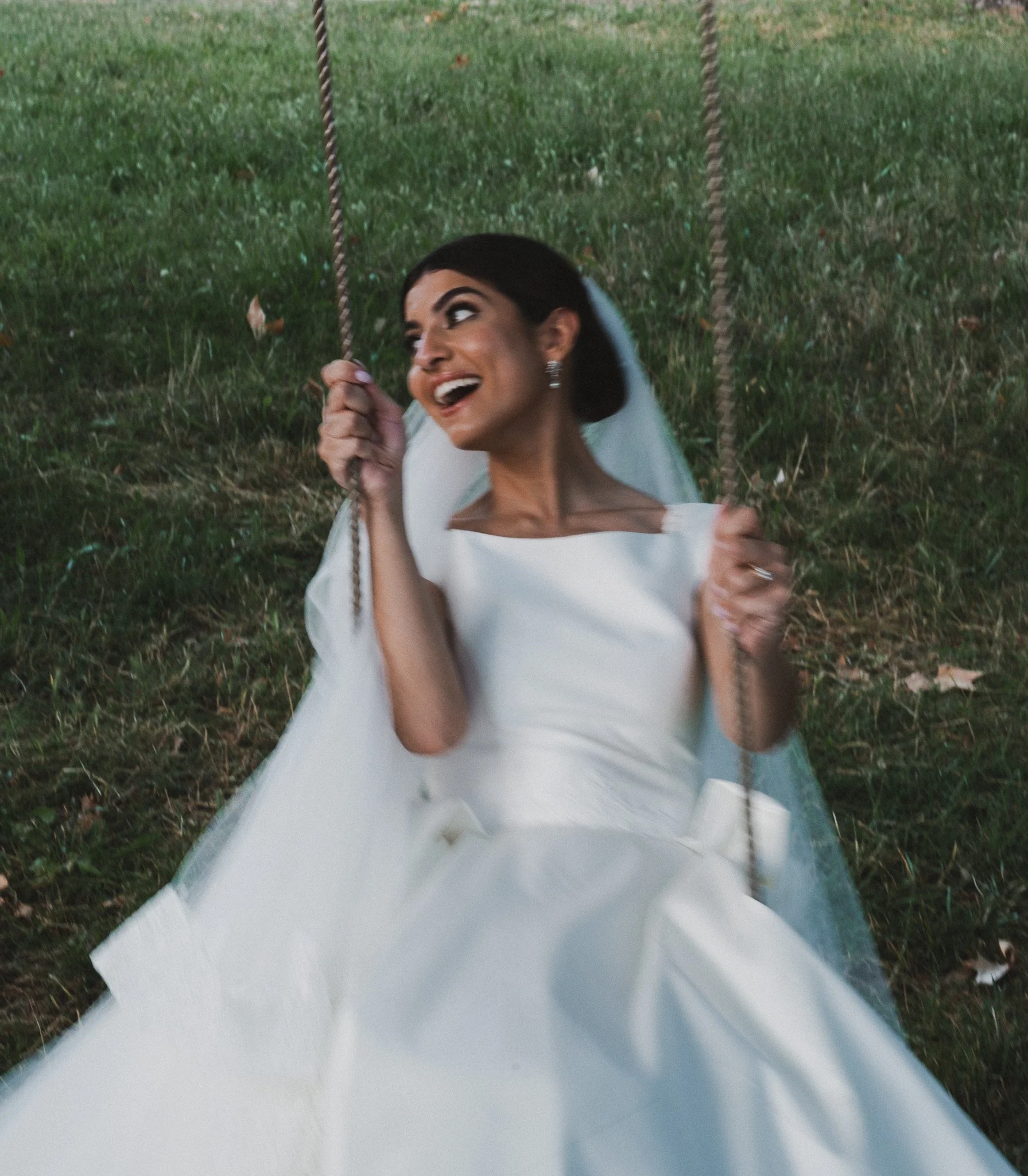 Femme en robe blanche riant sur une balançoire en plein air, entourée d'herbe verte et de feuilles tombées.