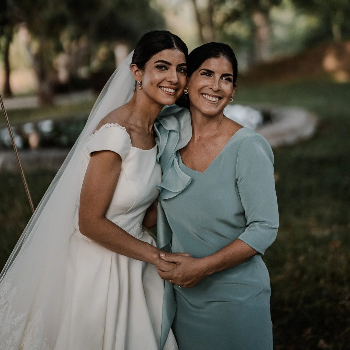 Deux femmes souriantes, dont une en robe de mariée et l'autre en robe bleue, se tiennent par la main dans un parc, lors d'une célébration de mariage