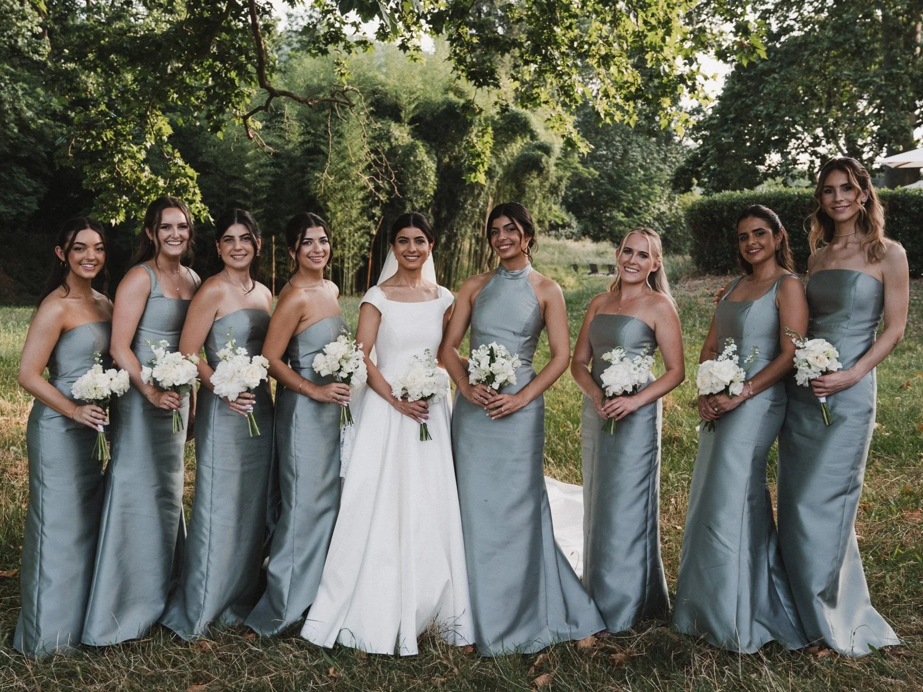 Groupe de neuf femmes, principalement bride et demoiselles d'honneur, posant à l'extérieur, dans un parc, lors d'un mariage, toutes tenant des bouquets de fleurs blanc.
