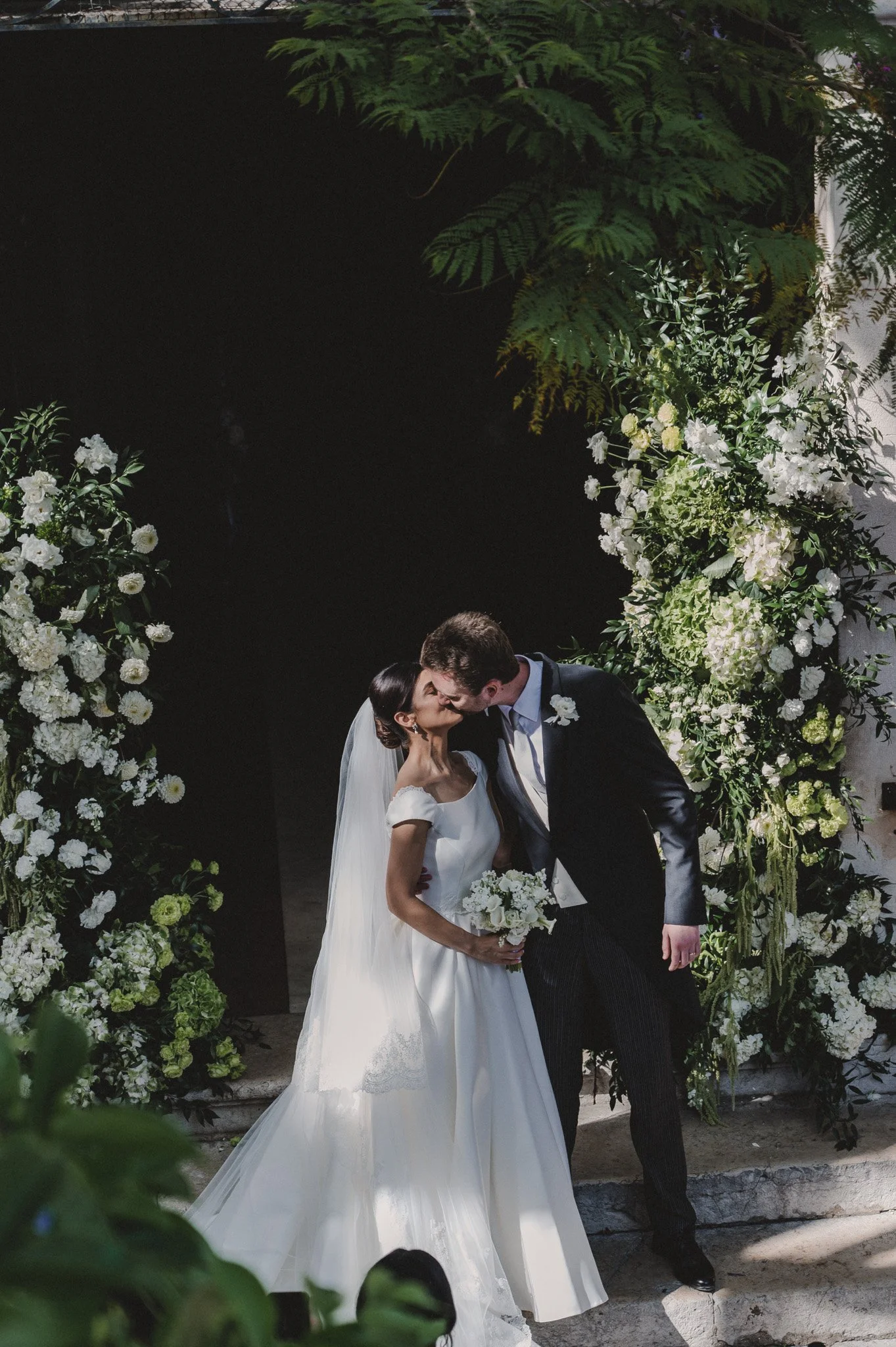 Un couple de mariés s'embrasse lors de leur mariage, entouré de décorations florales vertes et blanches, en extérieur, avec un fond sombre.