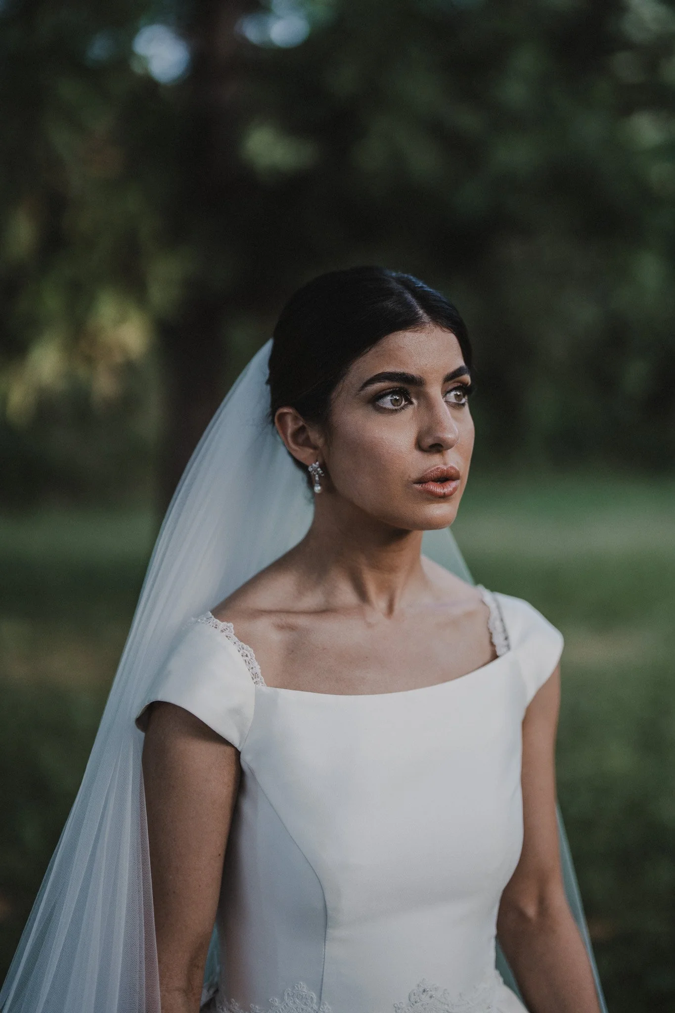 Une femme portant une robe de mariée blanche avec un voile, regardant vers la droite, avec un fond flou de nature.