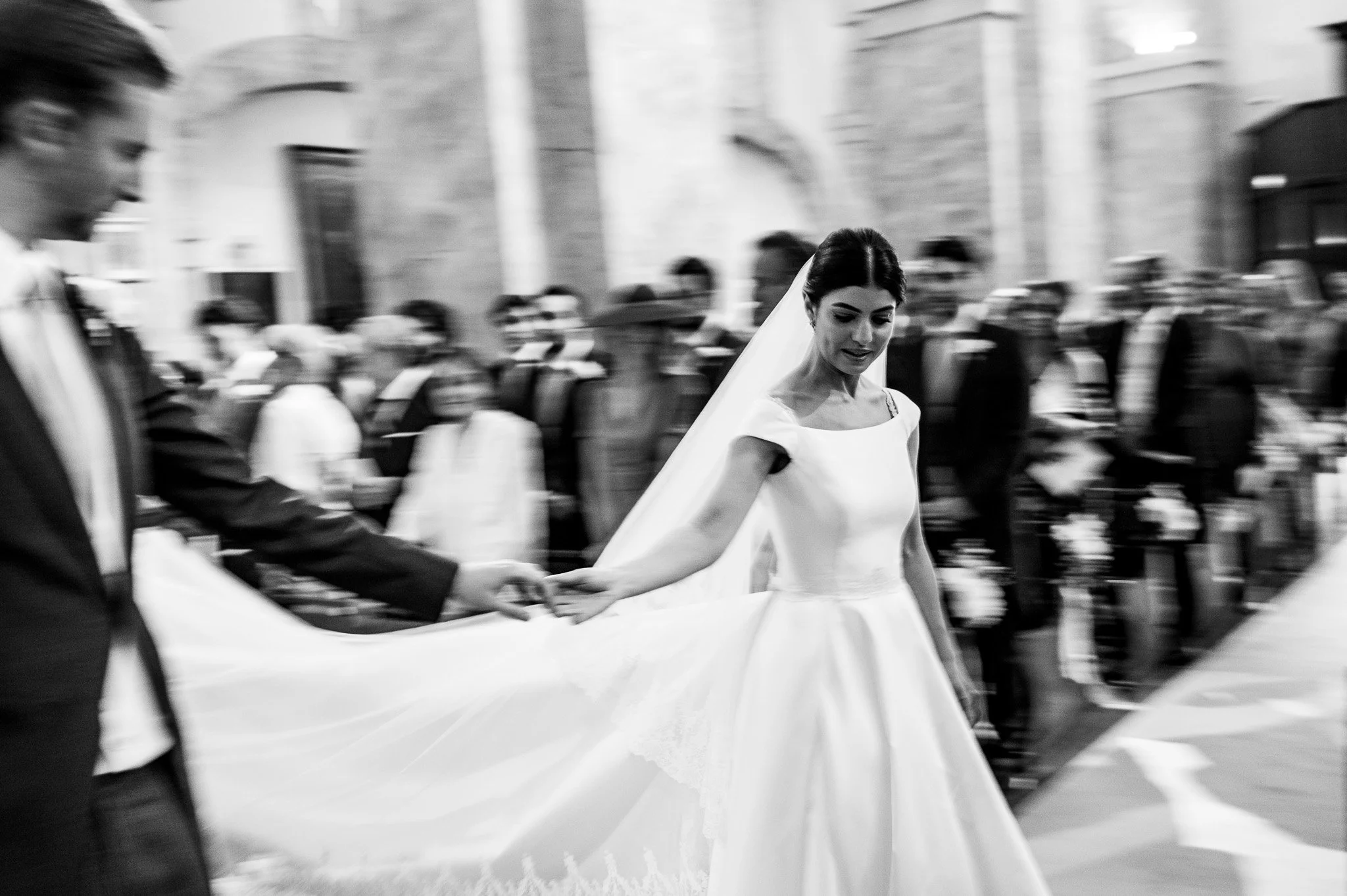 Une mariée portant une robe blanche et un voile, marchant dans une procession, entourée de personnes en costume, dans une église ou un lieu de cérémonie. En arrière-plan, il y a des silhouettes floues de personnes, renforçant l'effet de mouvement de l'image.