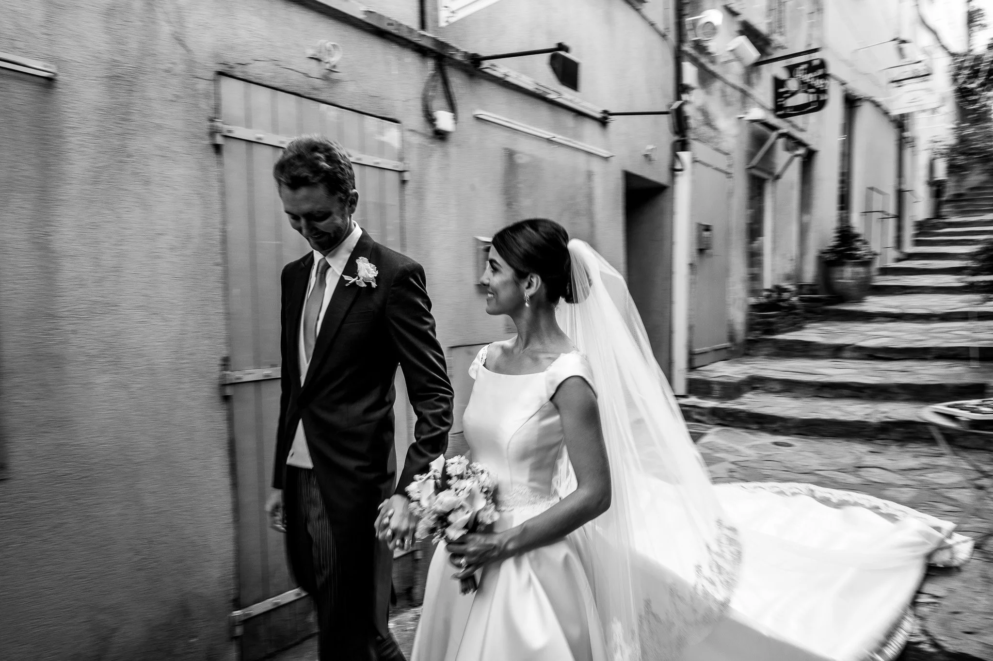 Un couple marié marche main dans la main dans une rue pavée, la femme porte une robe de mariée blanche avec un voile et tient un bouquet de fleurs, l'homme porte un costume sombre. La photo est en noir et blanc.