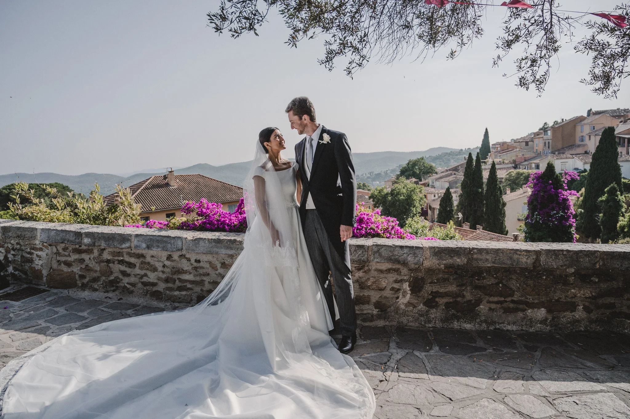 Un couple de mariés souriants se regarde tendrement en haut d'une terrasse avec des fleurs violettes et un paysage de collines en arrière-plan.