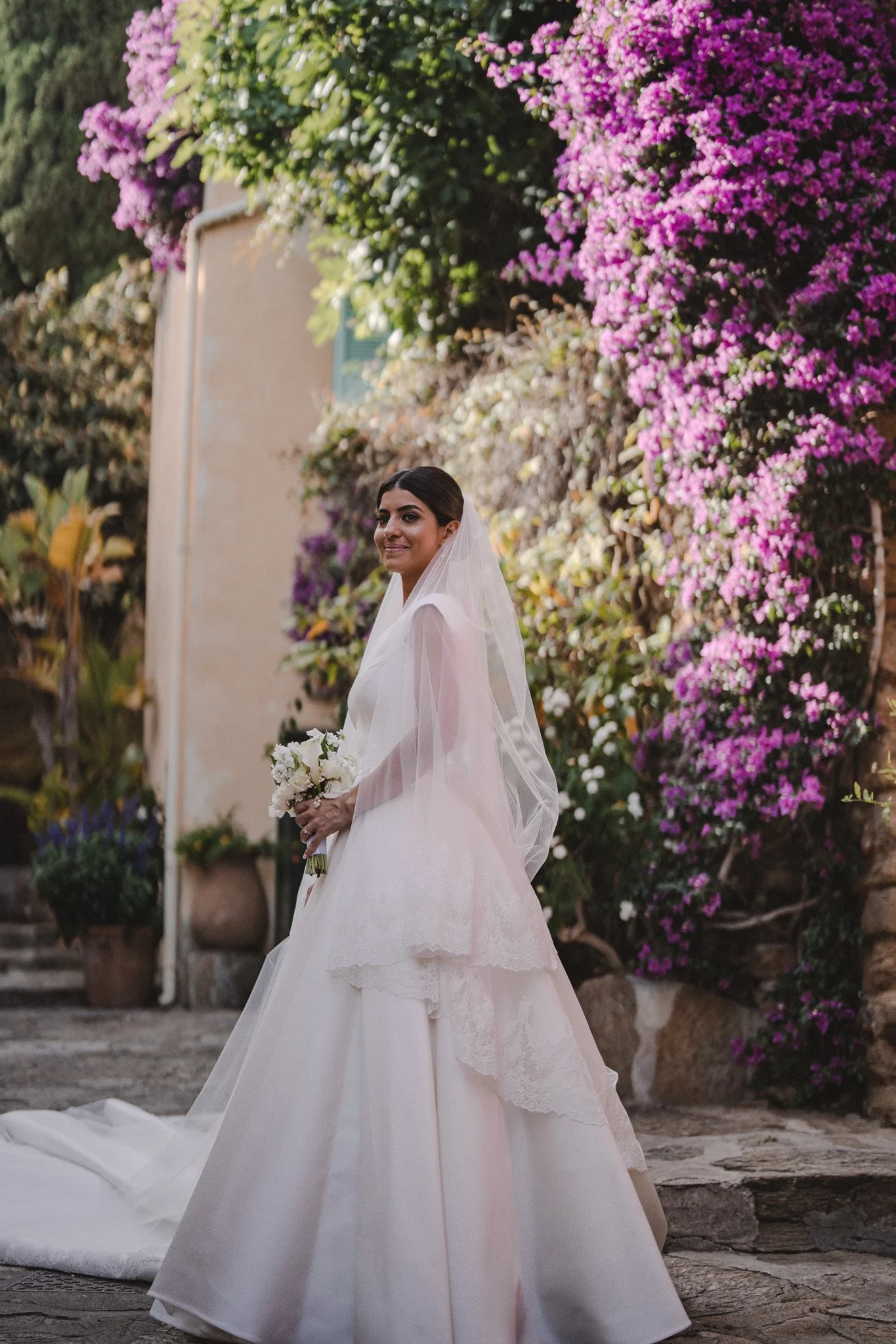 Femme en robe de mariage blanche, tenant un bouquet blanc, se tenant devant un mur couvert de bougainvilliers roses et violets.