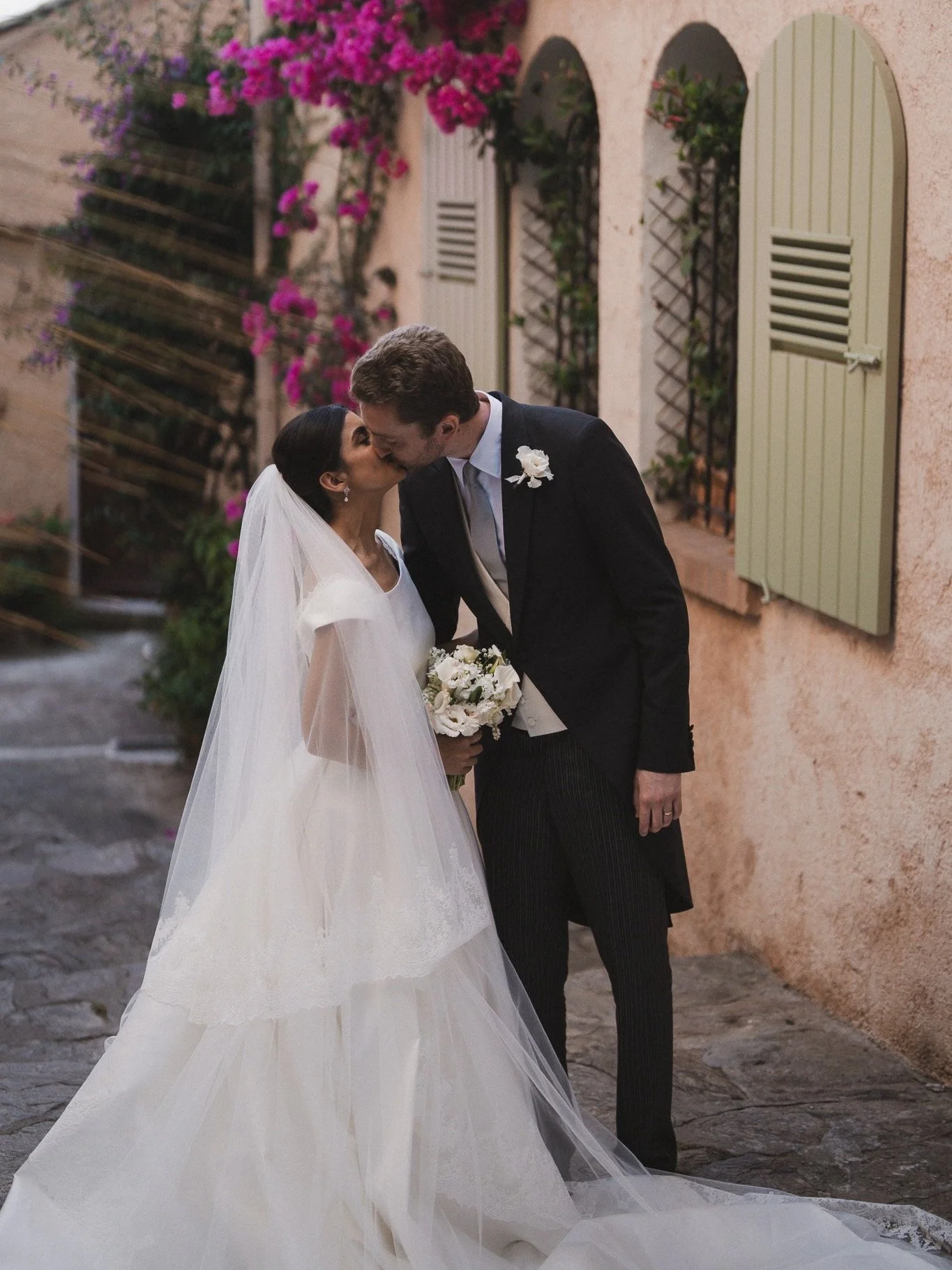 Un couple en robe de mariage et costume s'embrassent dans une rue pavée, ornée de volets verts et de bougainvilliers roses