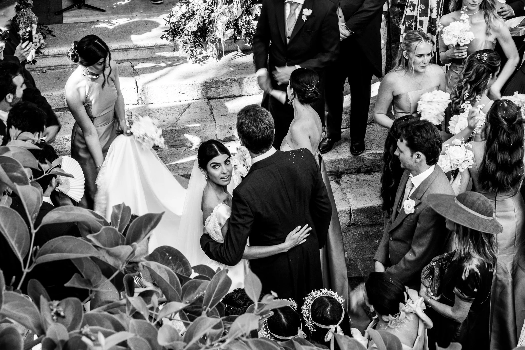 Un groupe de personnes lors d'un mariage, avec des femmes en robes de demoiselle d'honneur, un couple au centre, et d'autres invités portant des fleurs et des costumes, photographie en noir et blanc.