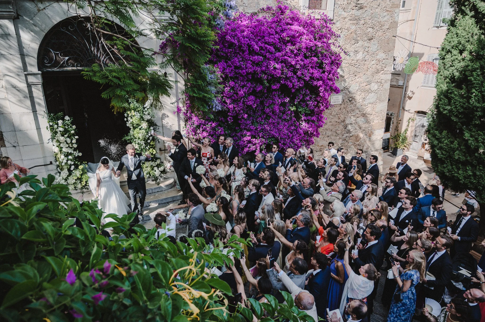 Un groupe de personnes en costume formel assistent à une mariage en extérieur, décorée avec des fleurs violettes et blanches, et un grand arbre avec des fleurs roses