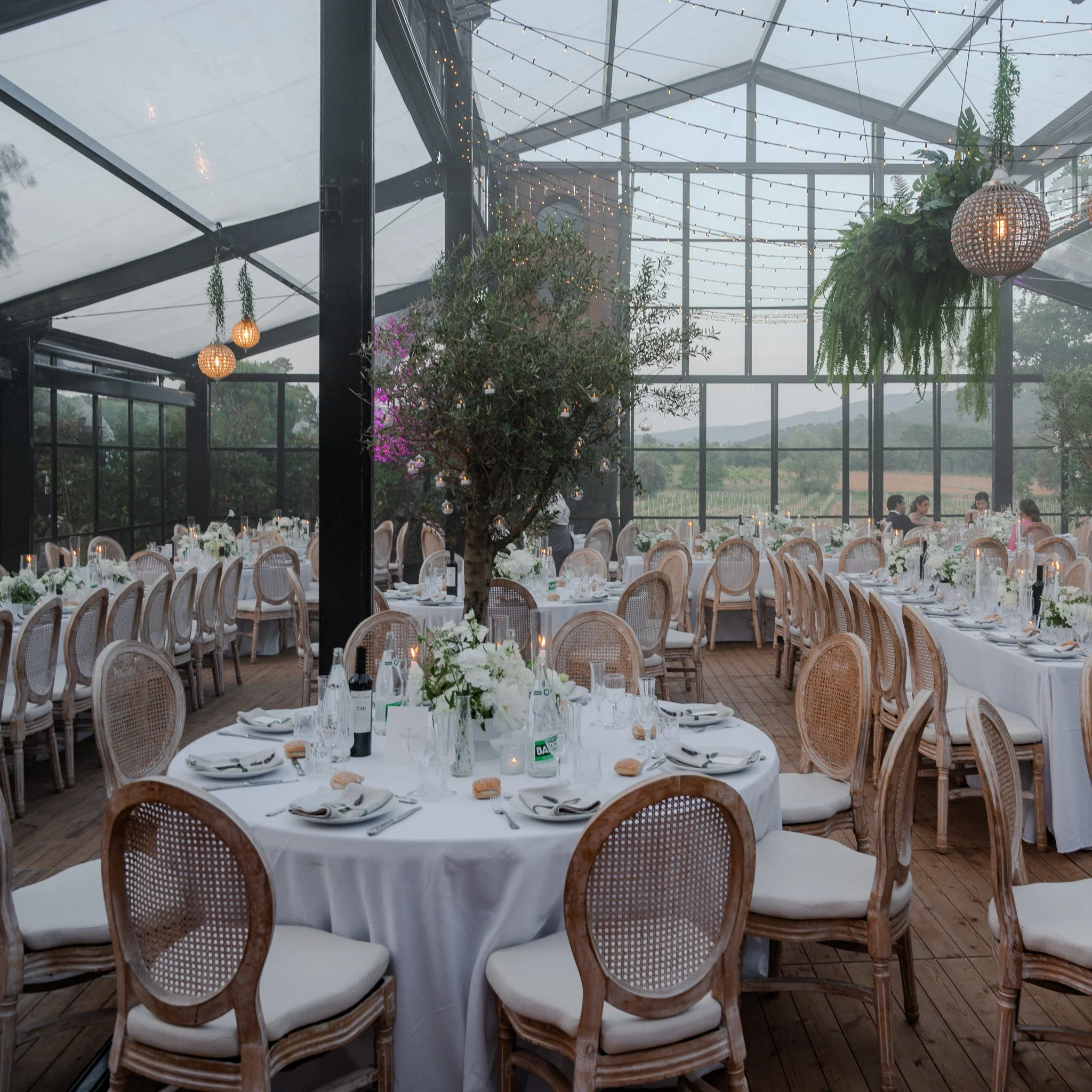 Salle de réception élégante avec des tables rondes couvertes de nappes blanches, décorées de bouquets de fleurs blanches, et entourées de chaises en bois au motif tressé, éclairée par des guirlandes lumineuses suspendues sous un toit en verre avec vue sur un paysage de campagne.