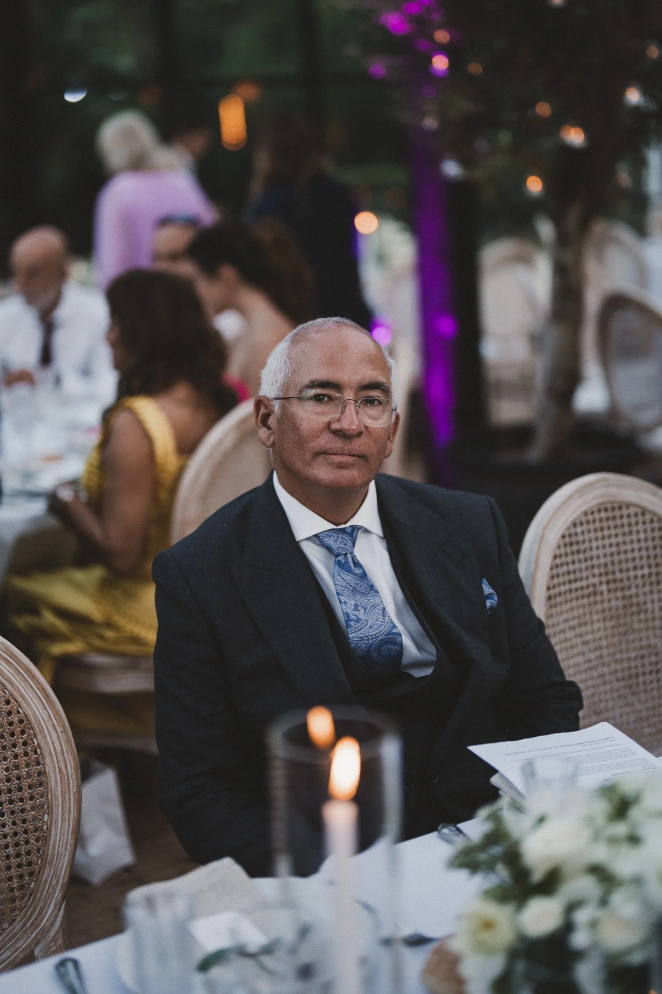 Un homme en costume noir et cravate assis à une table lors d'une cérémonie ou d'un repas, dans un lieu décoré avec des fleurs et une ambiance éclairée tamisée.