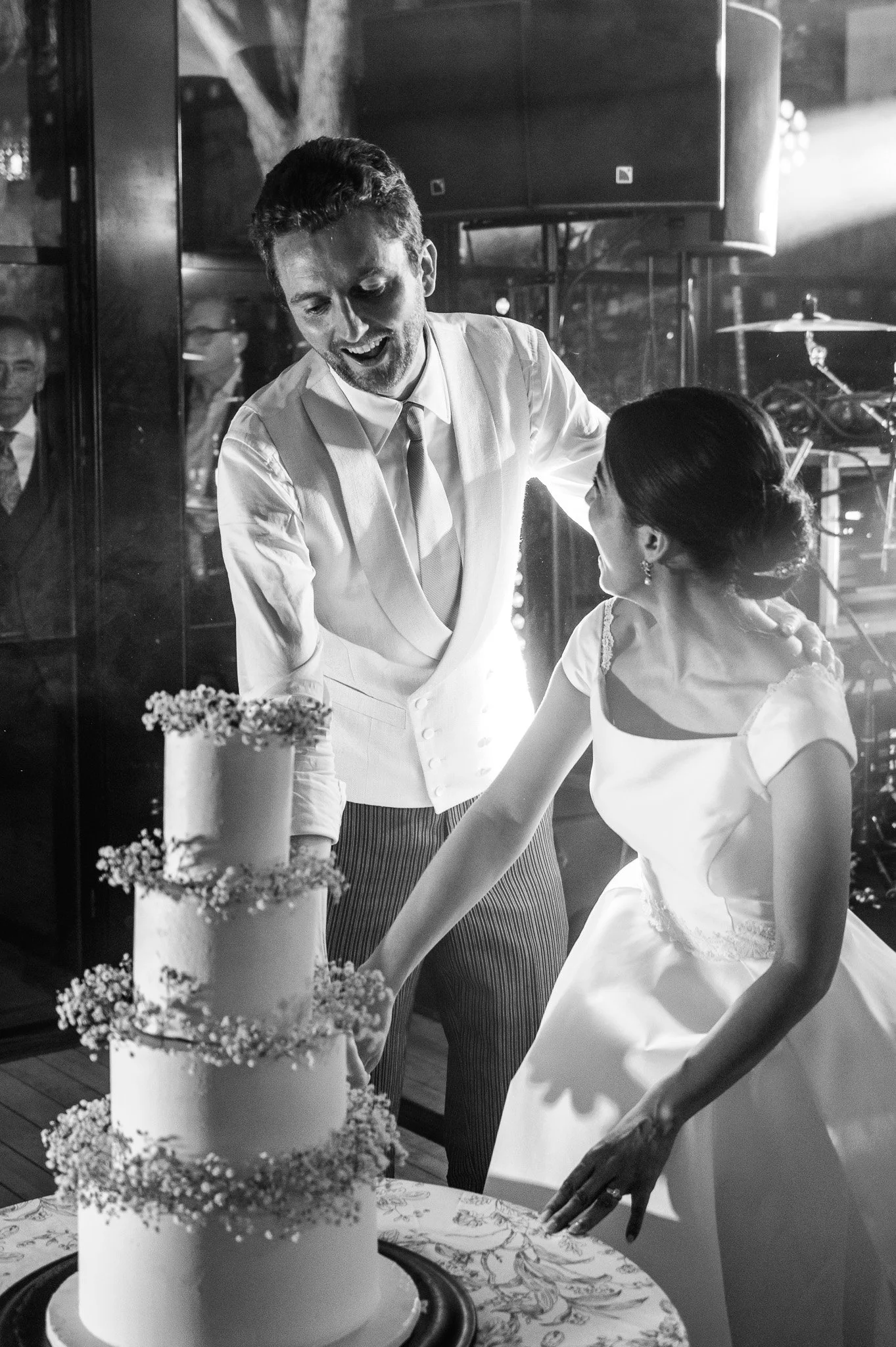 Un homme et une femme habillés en blanc coupent une pièce de gâteau lors d'une célébration, probablement un mariage, avec un gâteau à plusieurs couches décoré de fleurs.