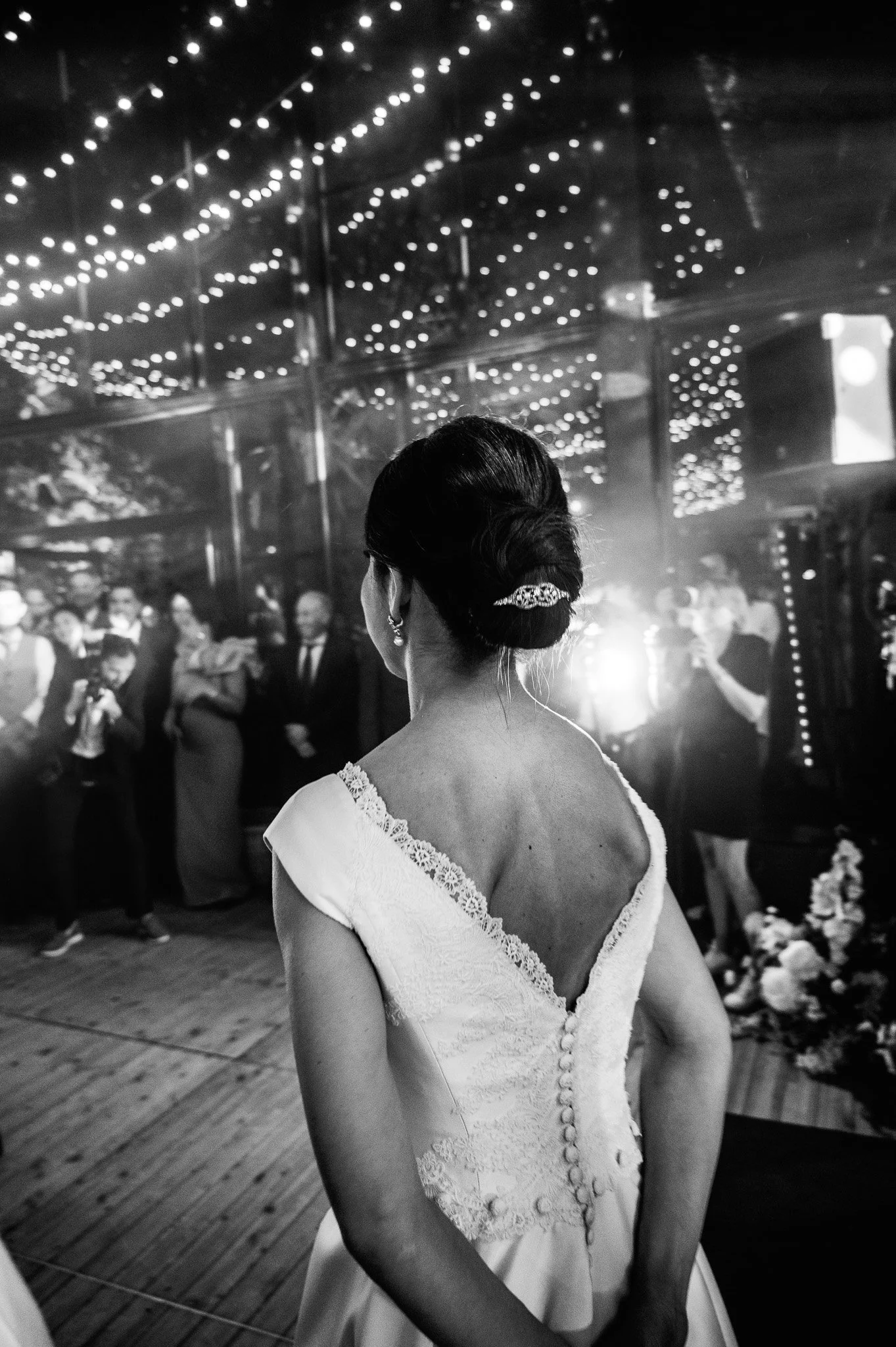 Une femme portant une robe de mariée vue de dos, dans une salle décorée de guirlandes lumineuses, lors d'une célébration ou un mariage.