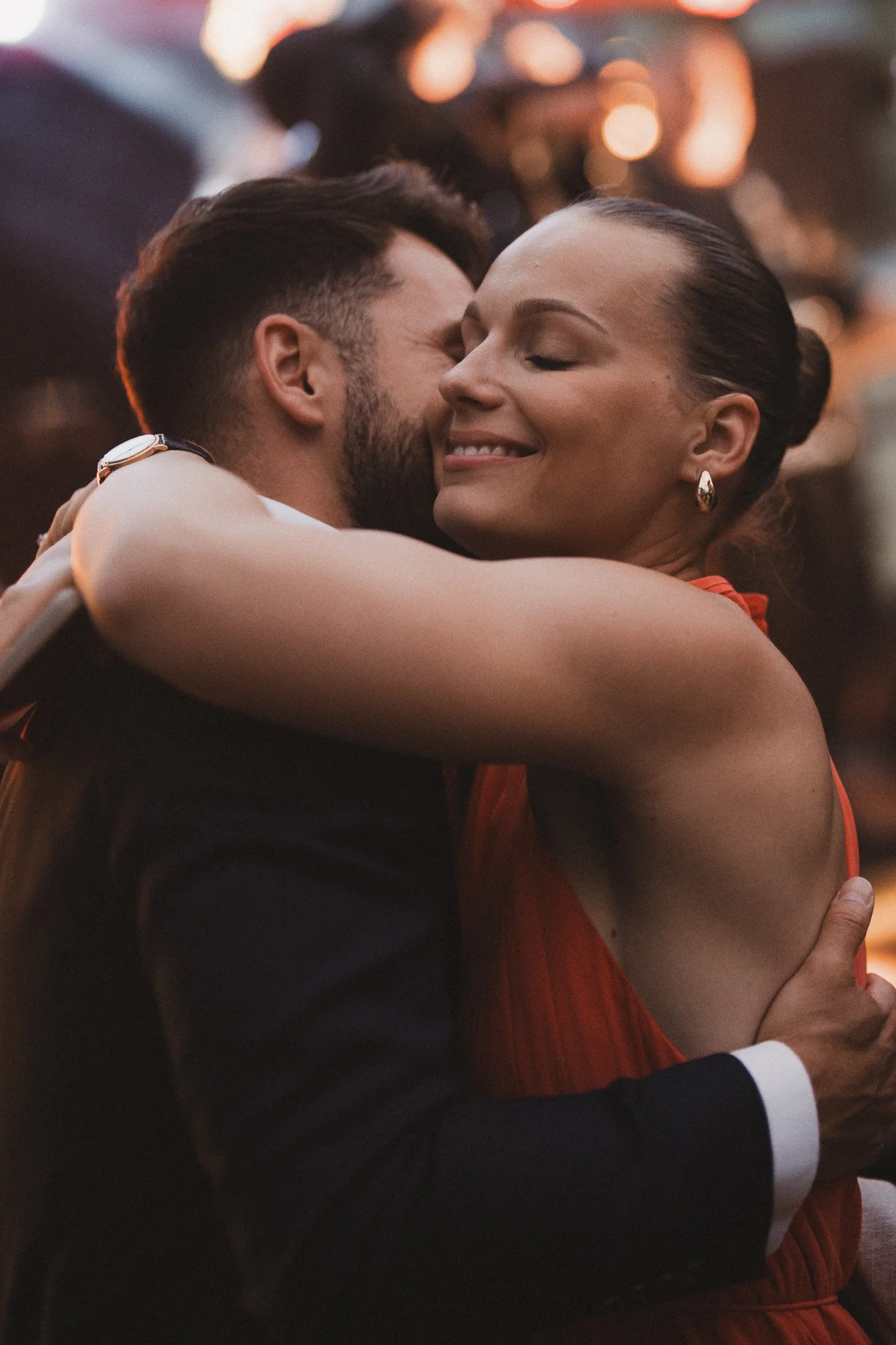 Un couple partage un câlin avec le regard fermé, exprimant de la tendresse, lors d'une soirée romantique dans un endroit éclairé par des lumières colorées.