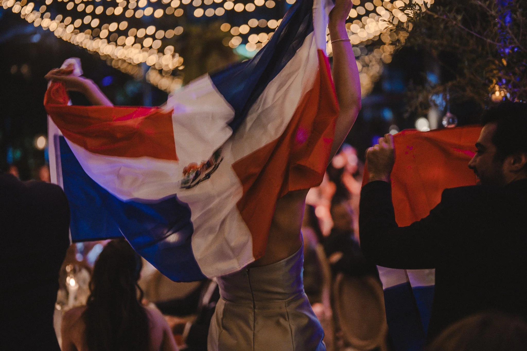 Des personnes dansent lors d'une fête en soirée, avec des guirlandes lumineuses en arrière-plan. L'une d'elles tient un drapeau français.