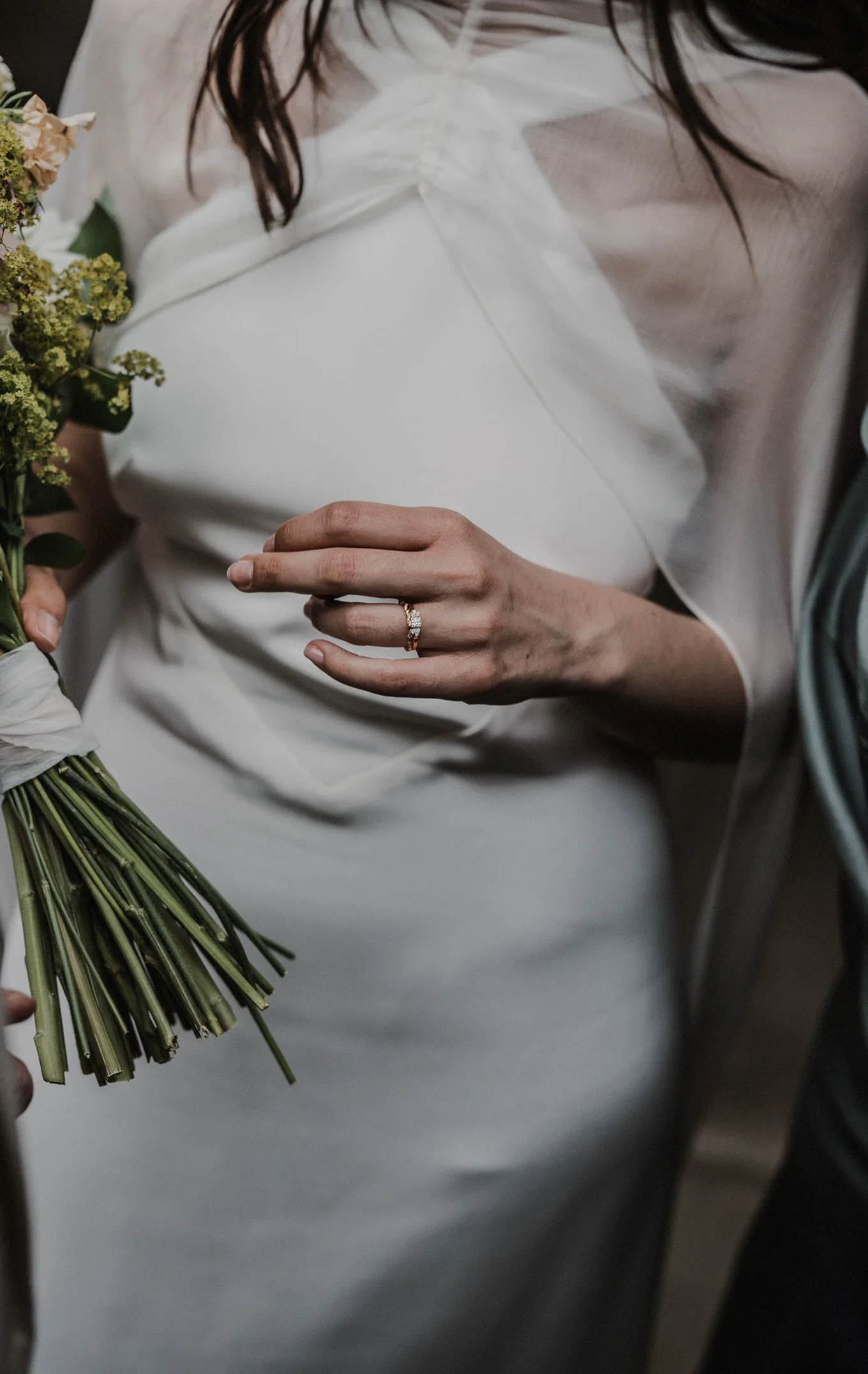 Femme tenant un bouquet de fleurs avec une main portant une alliance et une bague, vêtue d'une robe blanche en tissu doux, fond sombre.